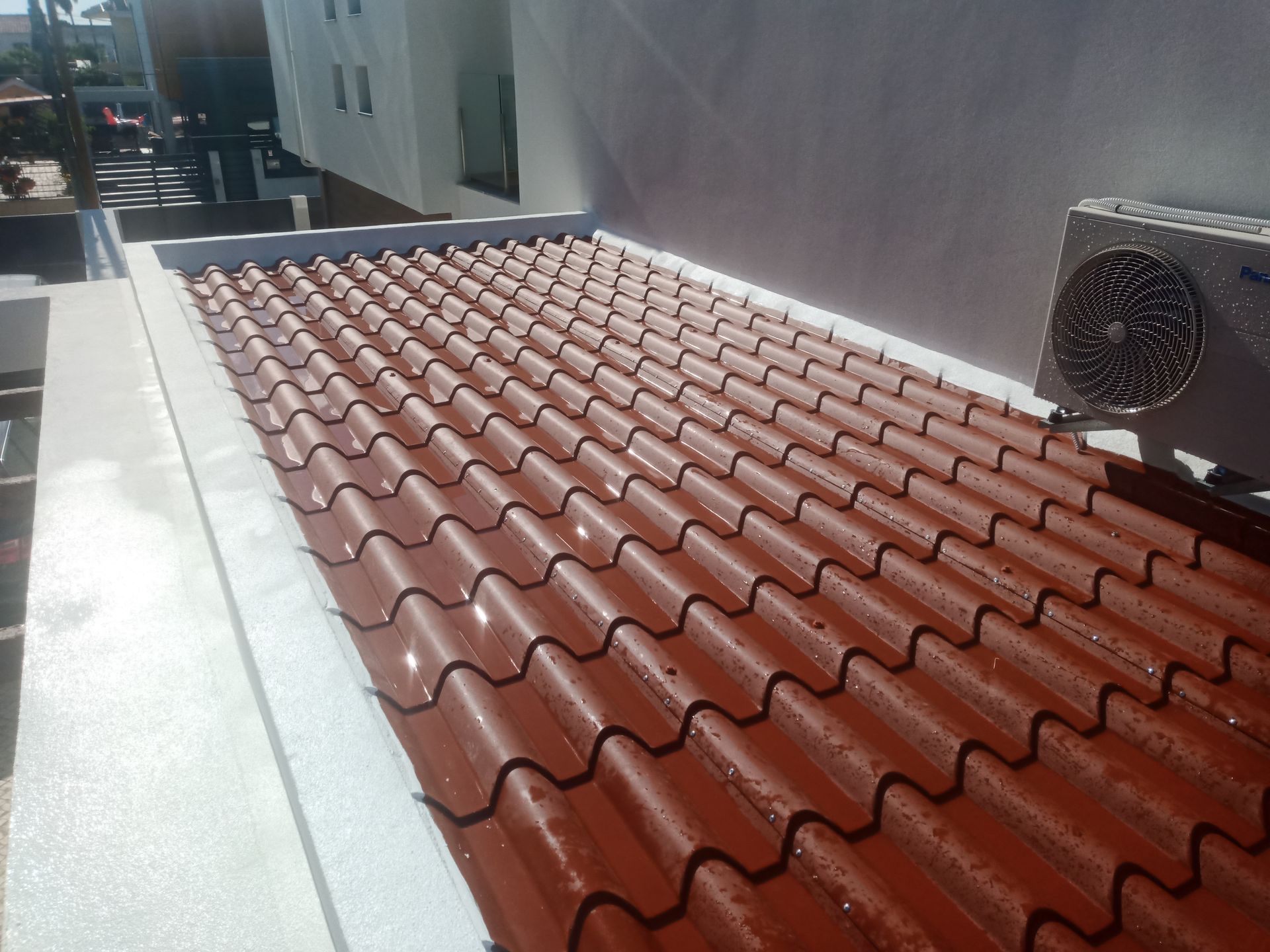 Telhado de telhas vermelhas onduladas com aspecto molhado, ao lado de uma parede branca e uma unidade de ar condicionado.