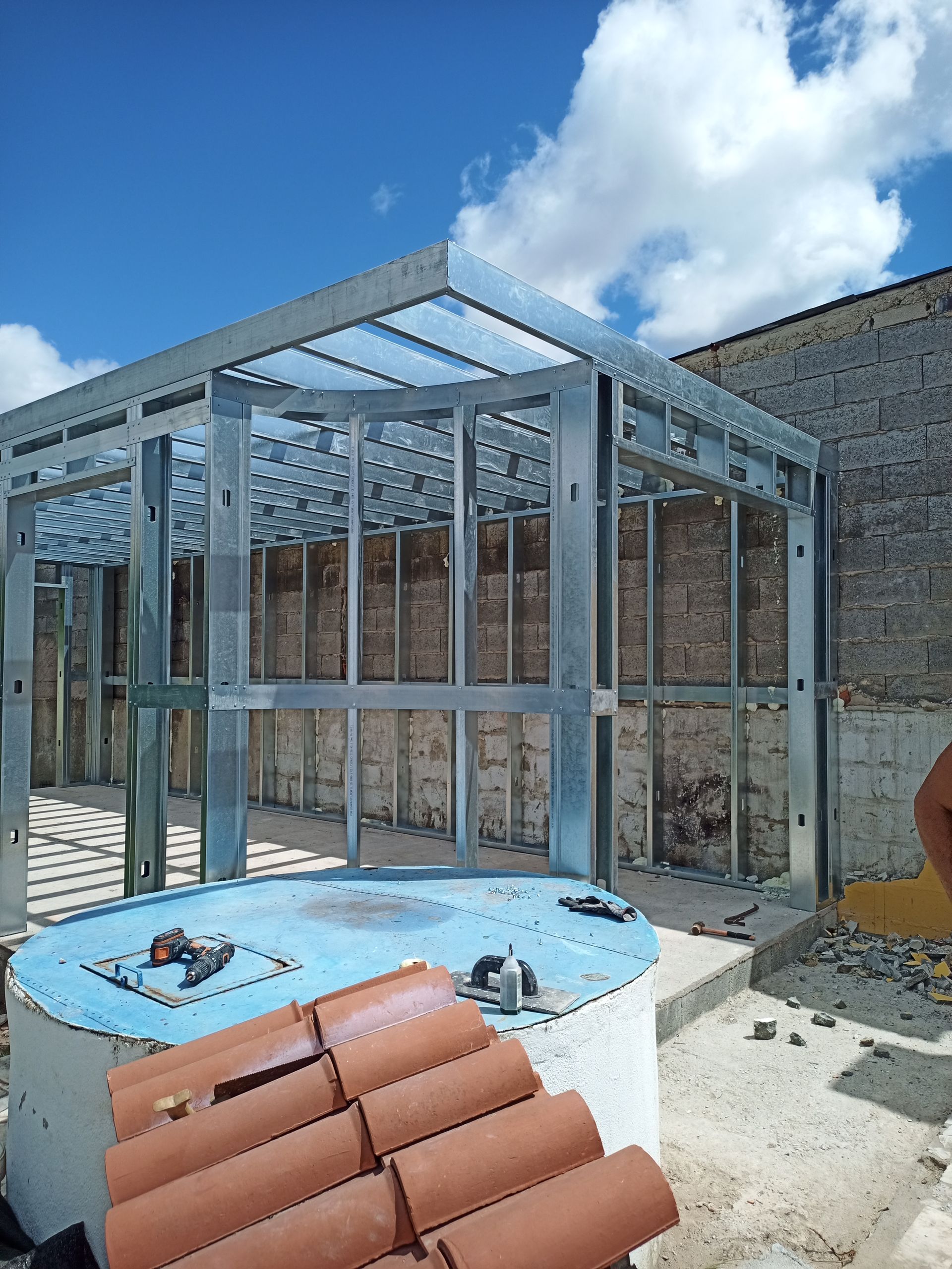 Estrutura metálica de um edifício com uma estrutura circular azul em primeiro plano, contra uma parede de tijolos e um céu claro.