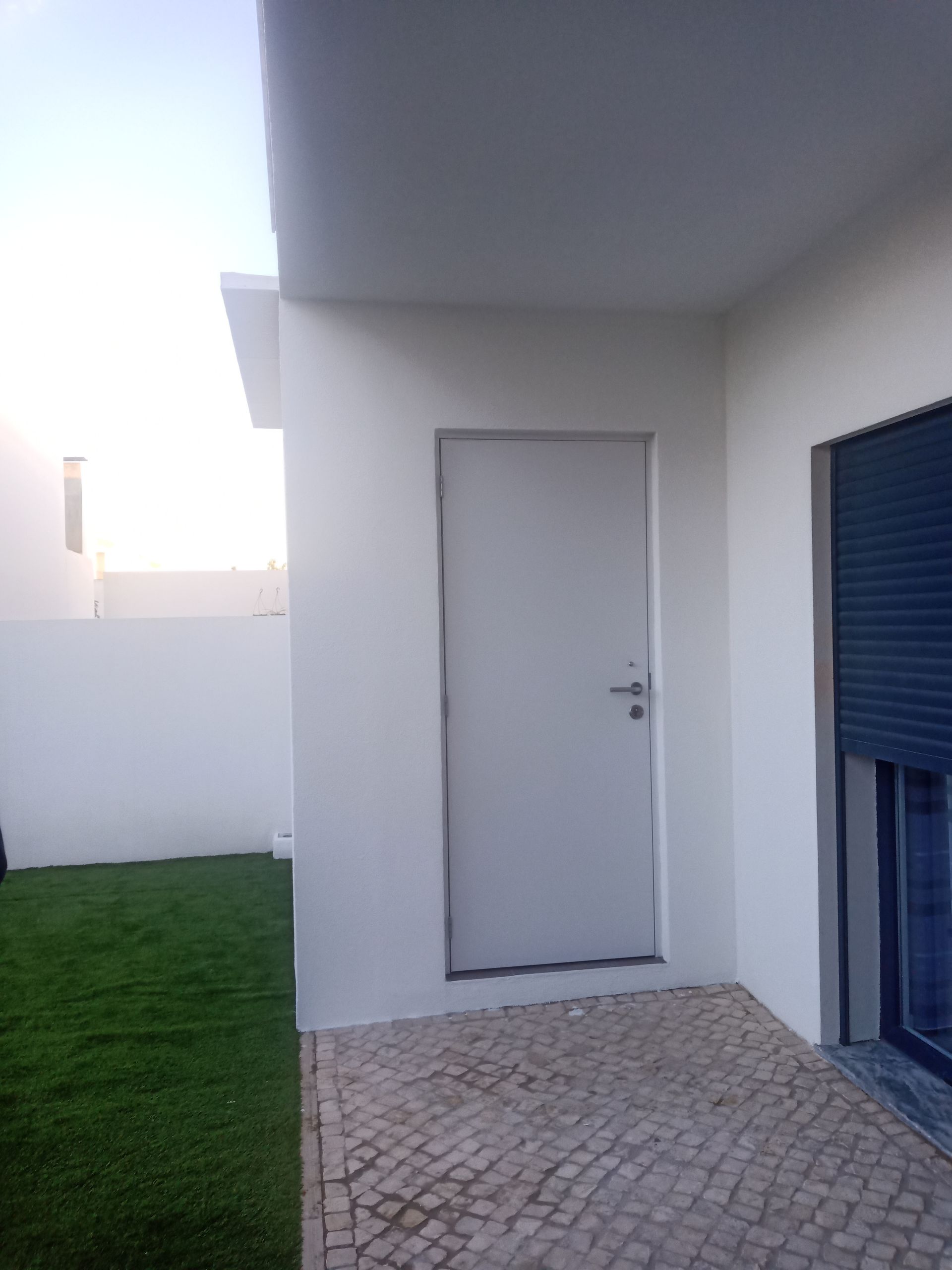 Porta branca na fachada branca de um prédio, ao lado de grama verde e uma janela com veneziana azul fechada.