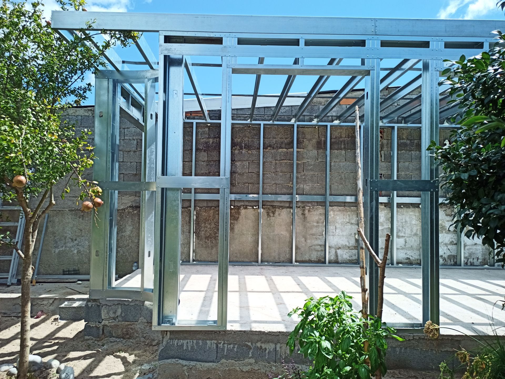 Estrutura metálica de um edifício em construção, ao ar livre, com uma parede de tijolos ao fundo.