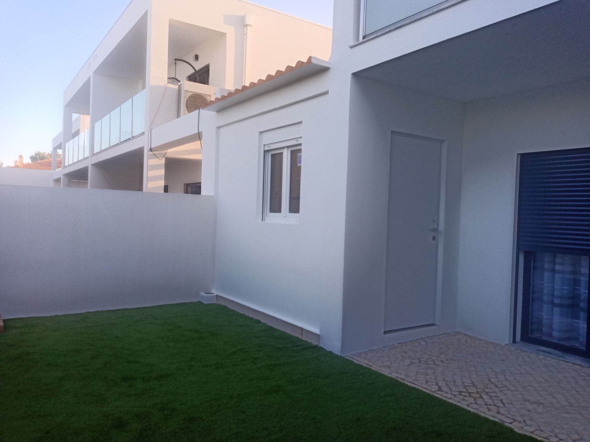 Vista exterior de um edifício branco com um pequeno galpão, grama verde e uma varanda com guarda-corpo de vidro.