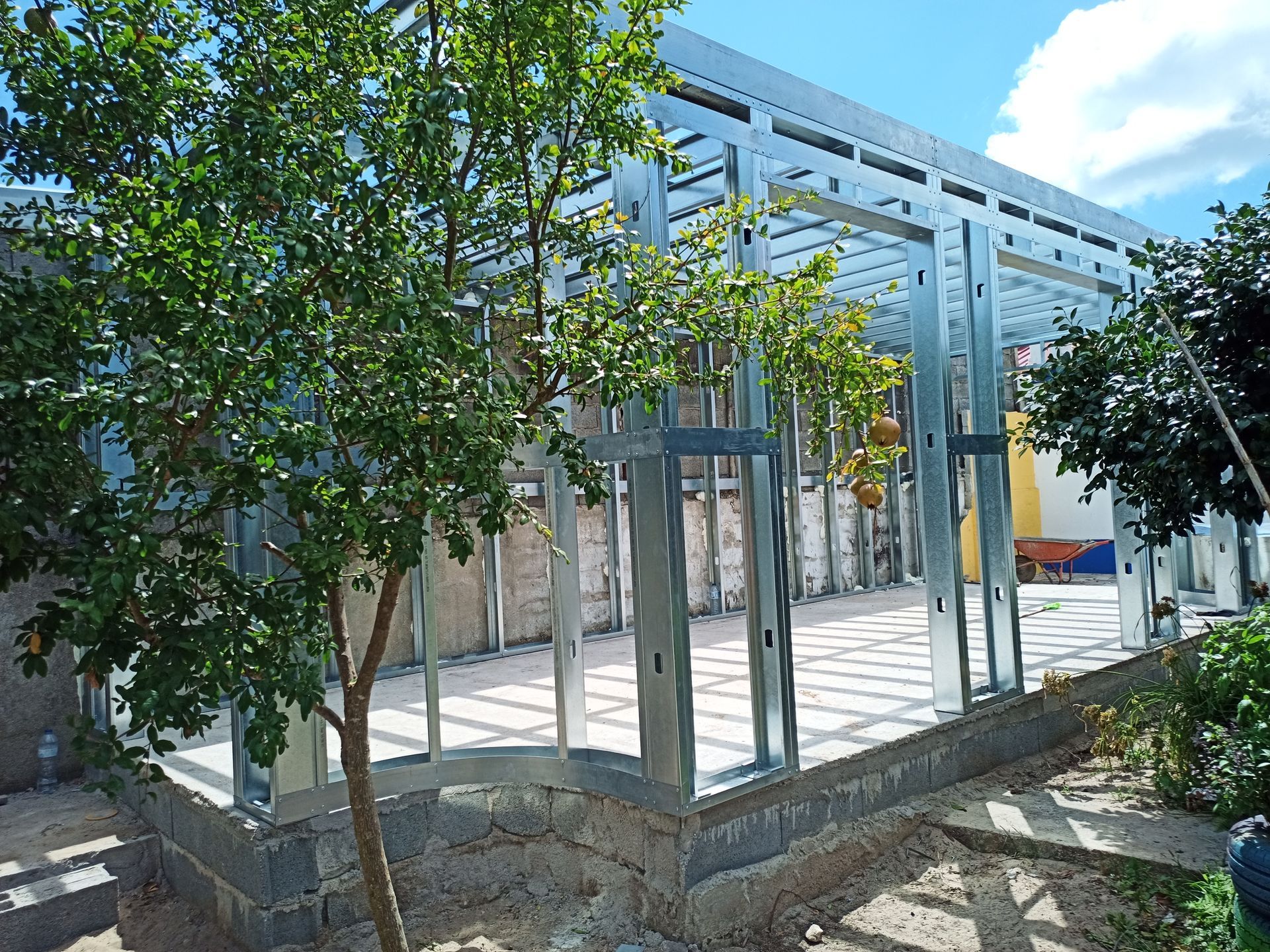 Estrutura metálica de um edifício em construção, com vista parcial de uma árvore verde em primeiro plano.