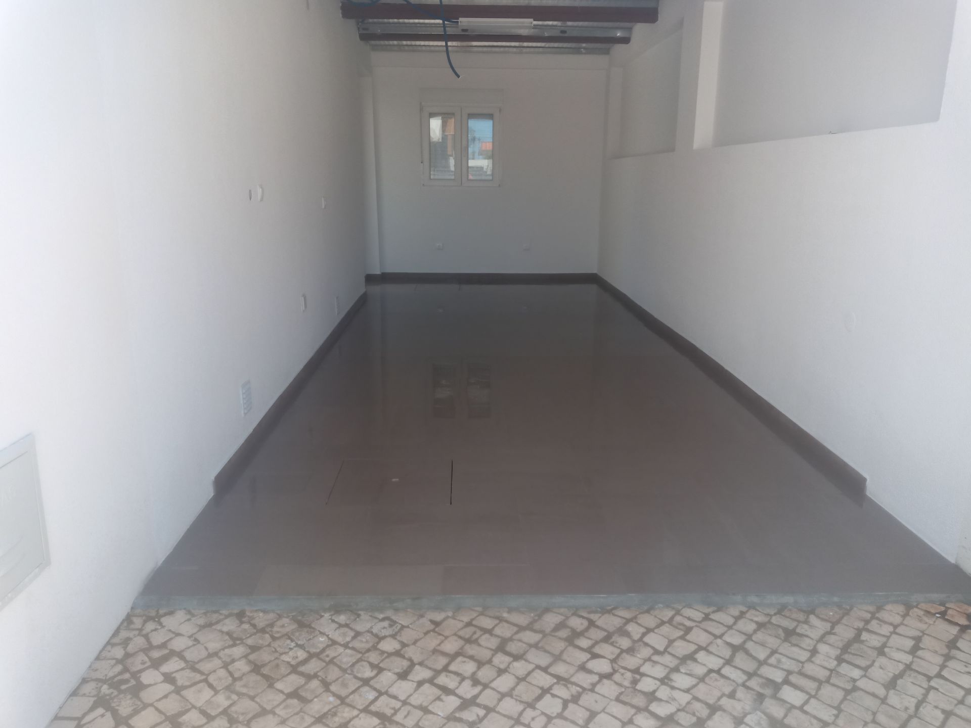 Quarto vazio, recém-construído, com piso liso e escuro e uma pequena janela ao fundo. Paredes brancas.