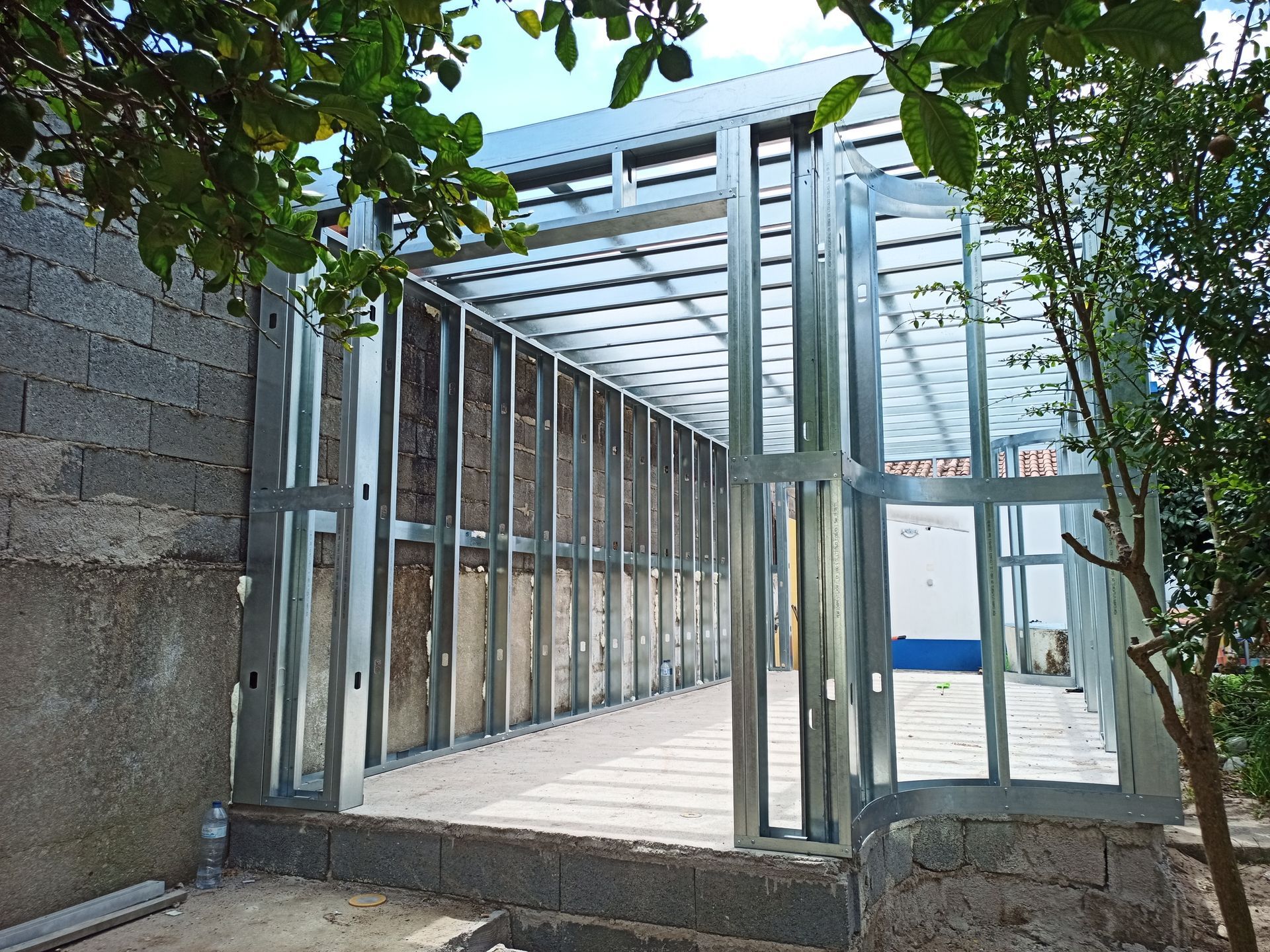 Estrutura metálica de uma construção em andamento, contra uma parede de blocos cinza. Porta aberta.