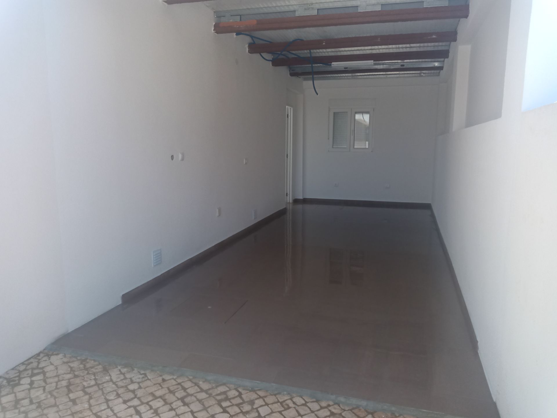 Sala comprida e vazia, com piso cinza e paredes brancas. Vigas de madeira expostas no teto e uma pequena janela ao fundo.