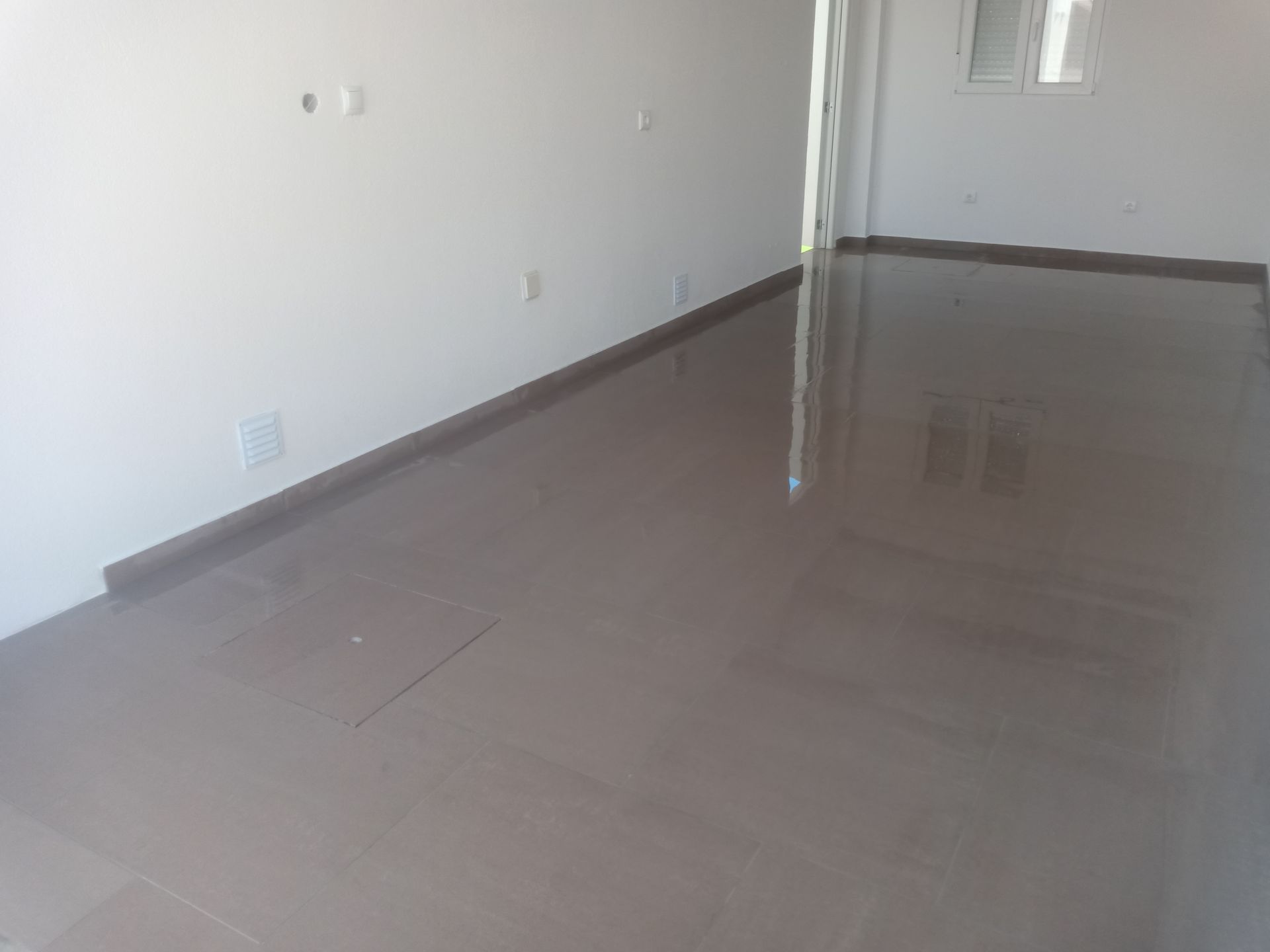 Sala vazia com piso marrom e paredes brancas; uma fonte de luz reflete no piso brilhante.