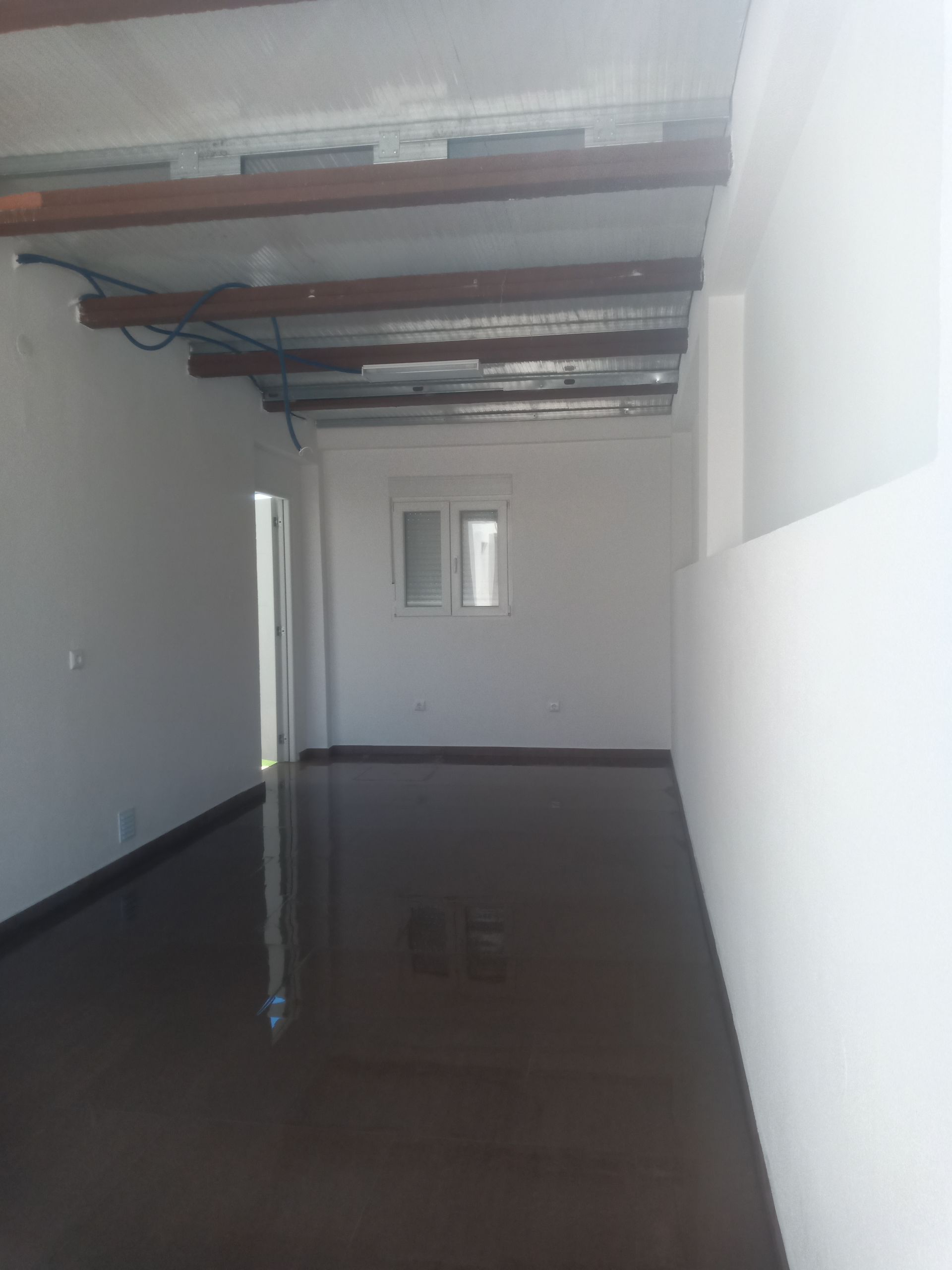 Sala retangular vazia com piso marrom, paredes brancas e vigas de madeira expostas no teto.