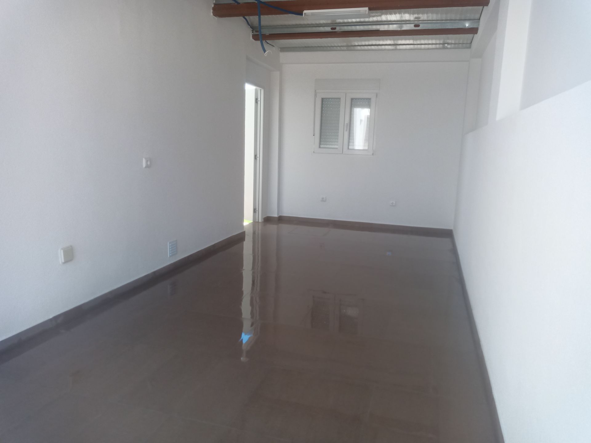 Quarto vazio com piso marrom brilhante, paredes brancas, janela pequena e porta aberta.