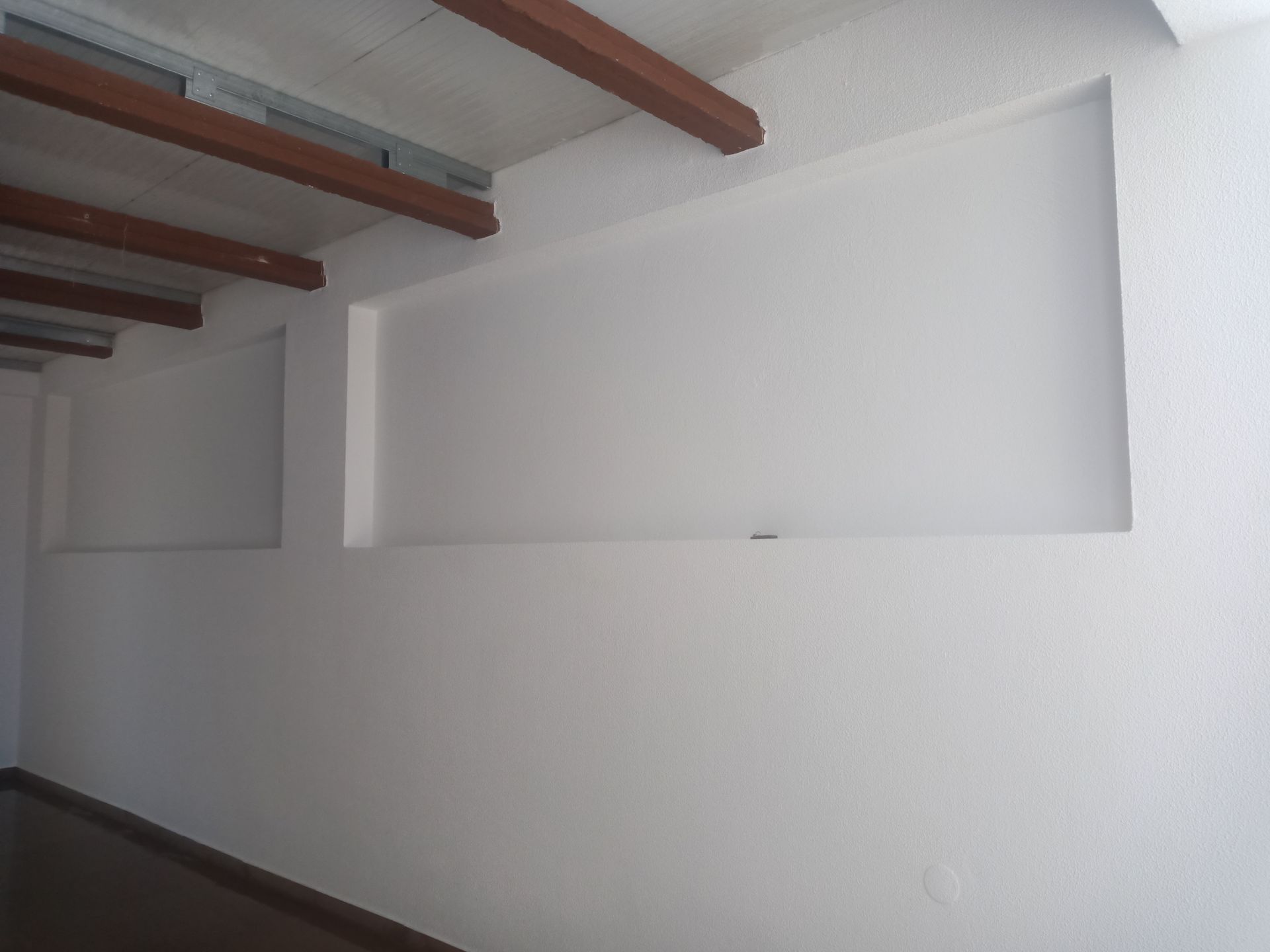 Parede interna branca com nichos retangulares e vigas de madeira no teto.