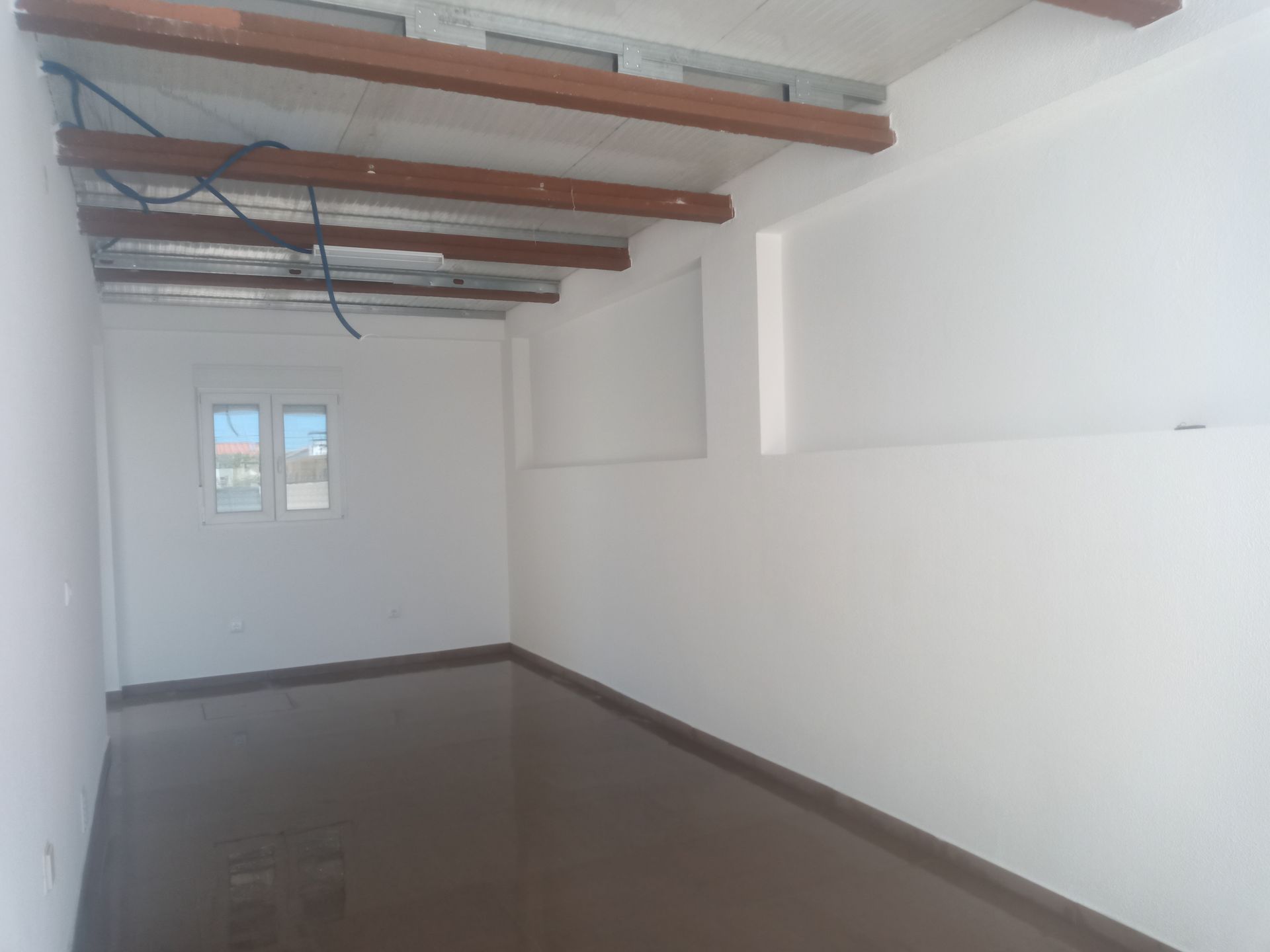 Quarto vazio com paredes brancas, janela, vigas escuras e piso marrom brilhante.