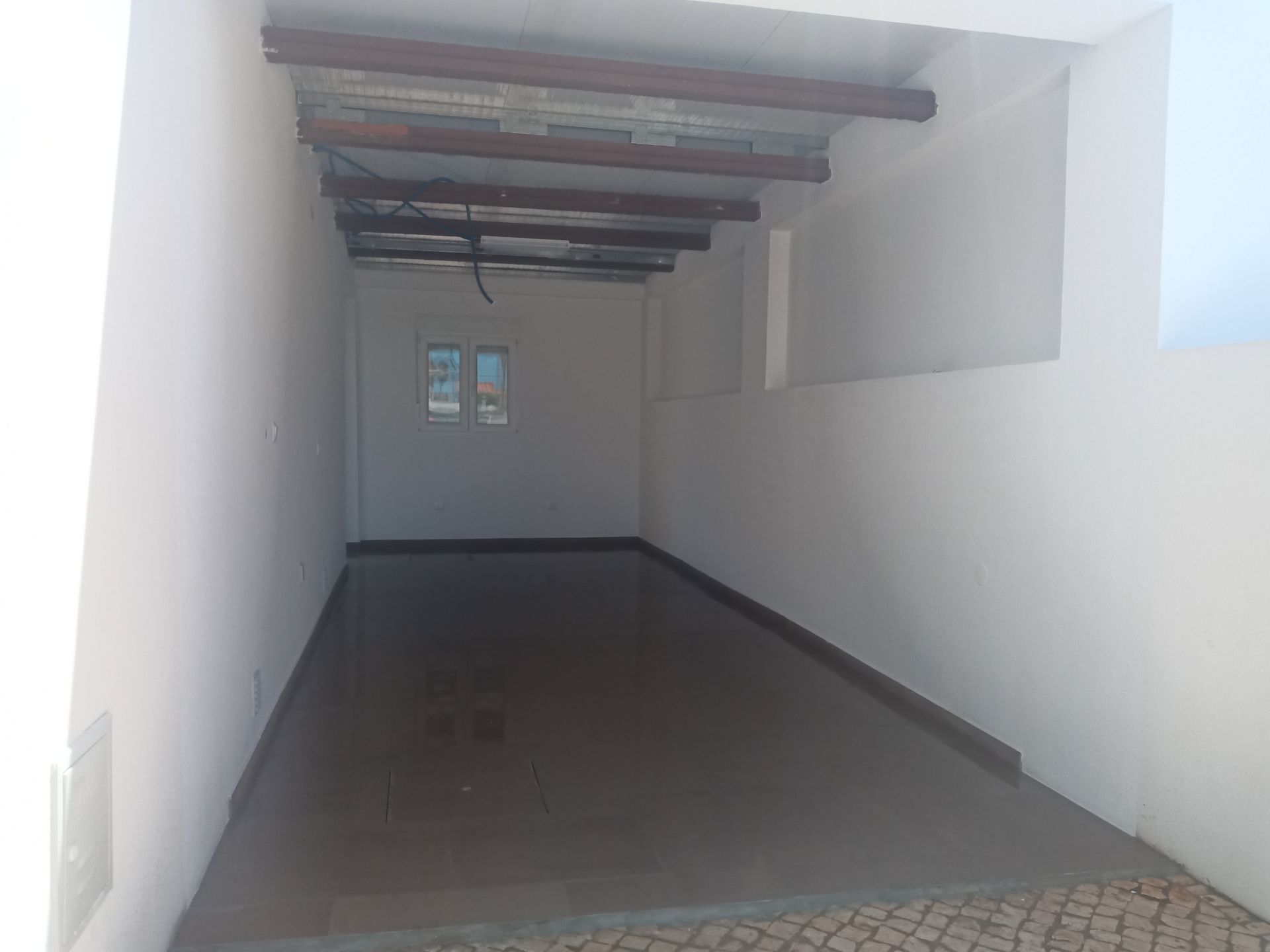 Beco estreito, com paredes brancas, piso de concreto, vigas de madeira no teto e uma pequena janela nos fundos.