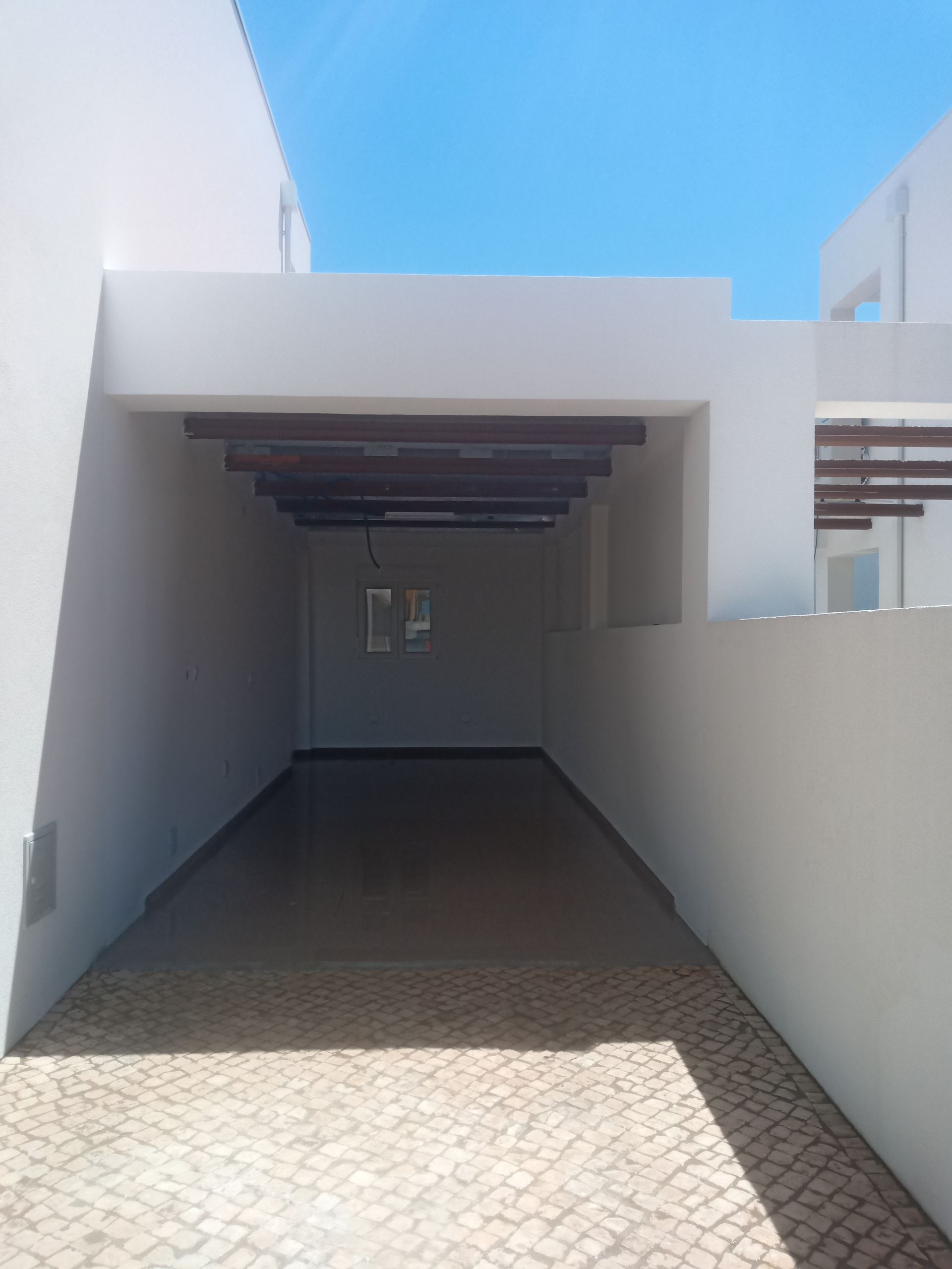 Passarela coberta com vigas de madeira escura, paredes brancas e caminho de paralelepípedos em um dia ensolarado.