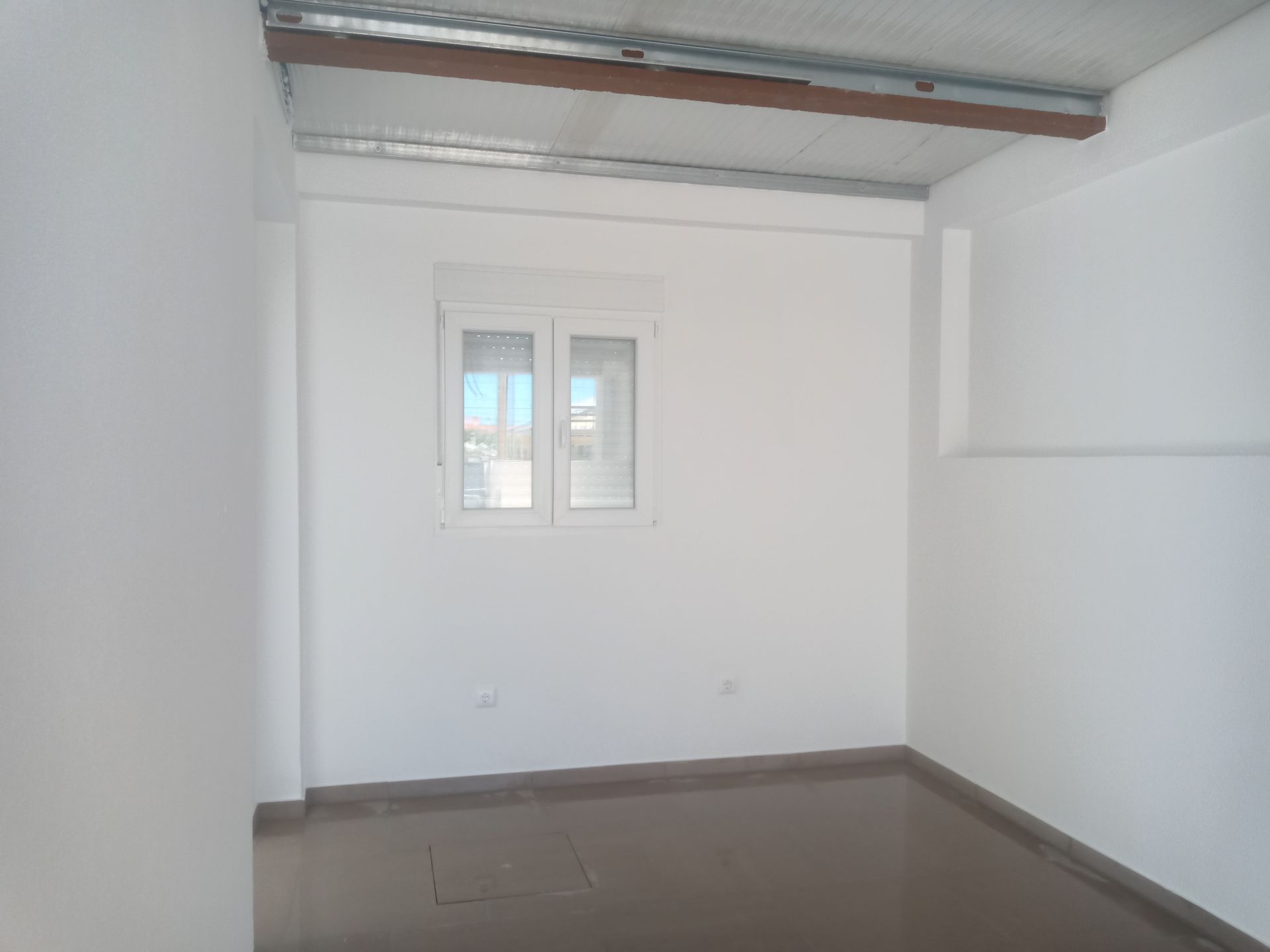 Quarto branco vazio com uma pequena janela, paredes nuas e uma viga visível no teto.