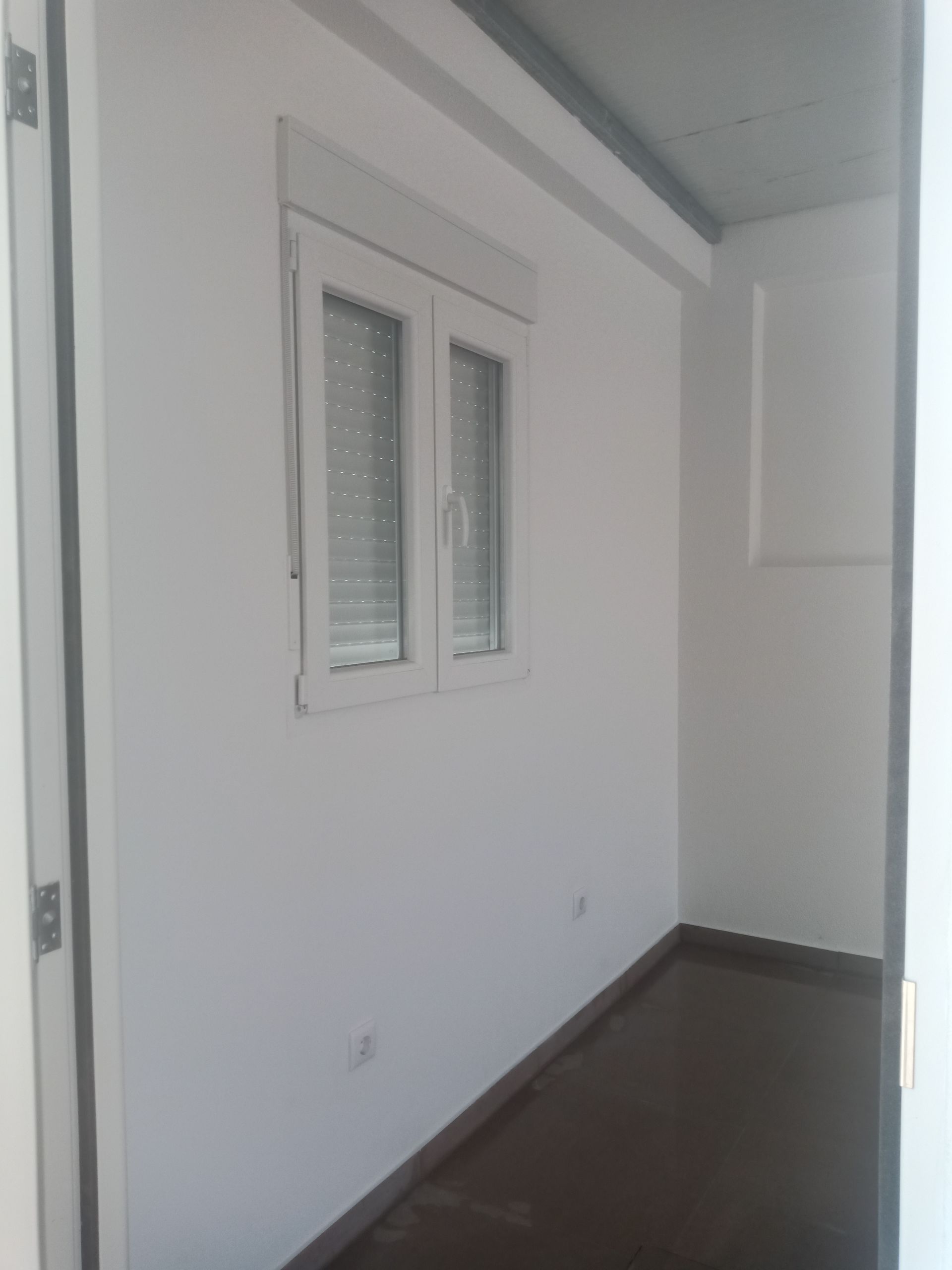 Quarto com paredes brancas e janela com persianas fechadas. Piso escuro e batente de porta à direita.