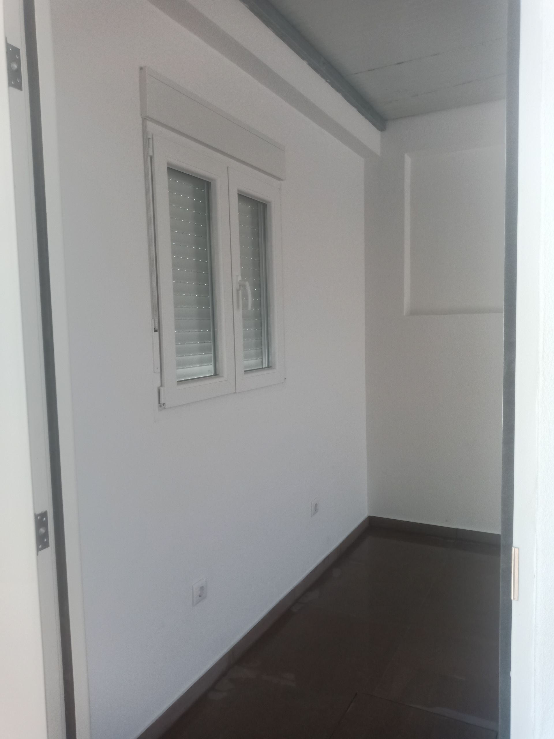 Um quarto vazio com paredes brancas, uma janela, chão escuro e uma porta aberta à direita.