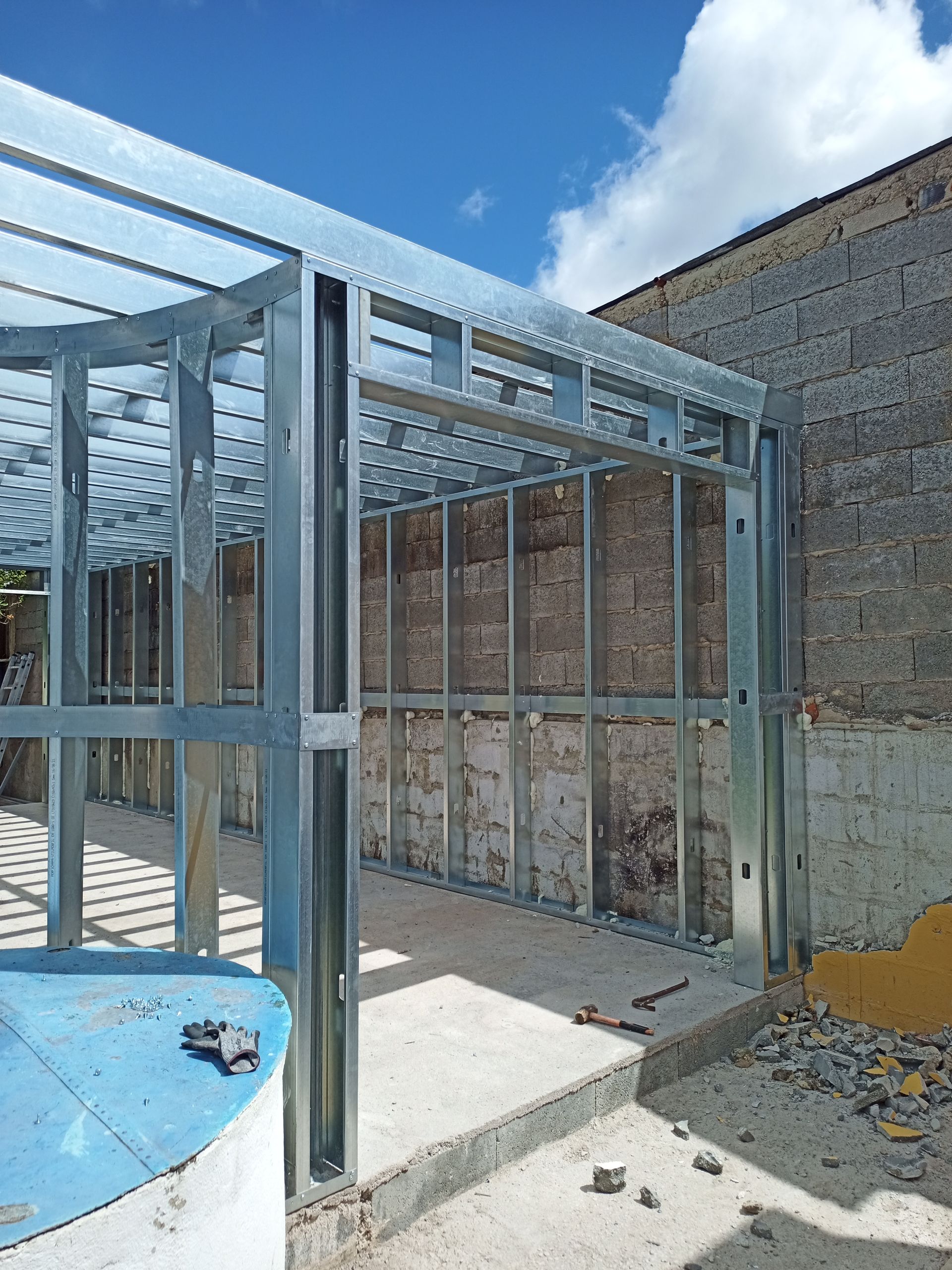 Estrutura metálica de uma construção em andamento, com piso de concreto e parede de tijolos ao fundo.