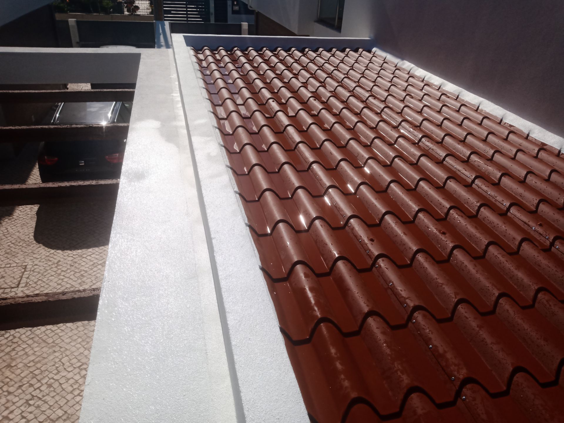 Telhado de zinco vermelho ao lado de uma parede branca, visto de cima. A luz do sol brilha nas telhas.
