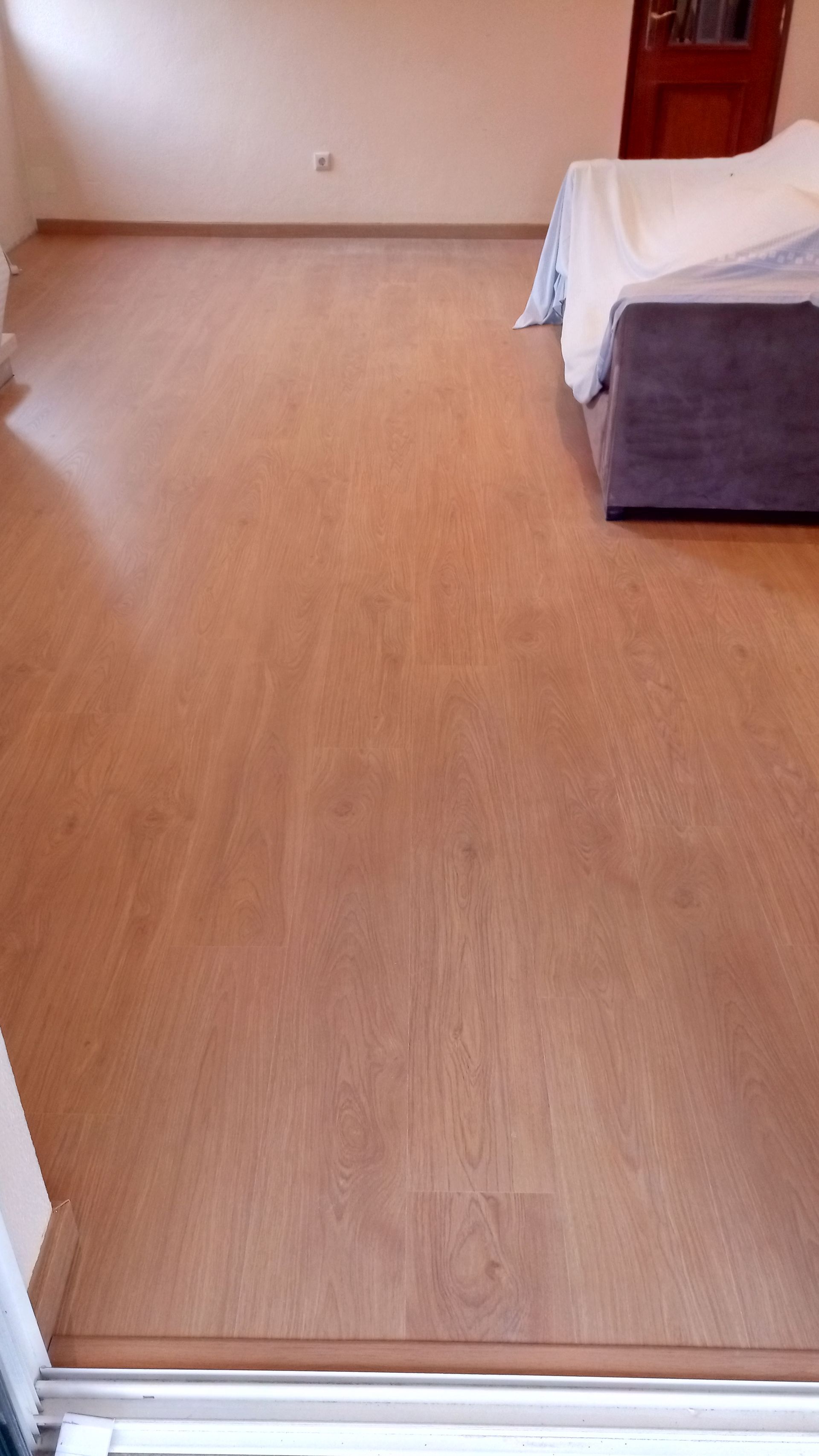 Piso de madeira em um quarto, com um sofá parcialmente coberto e uma porta ao fundo.