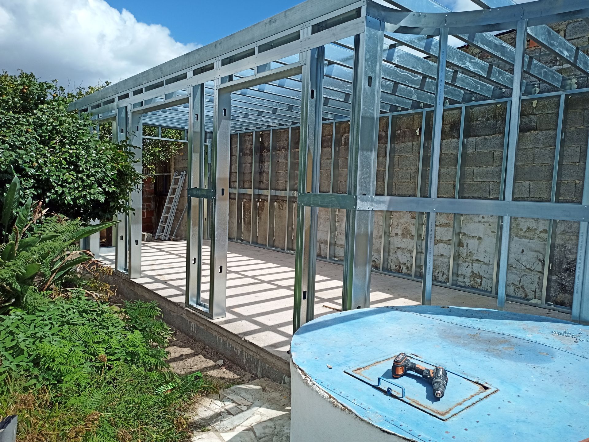 Estrutura de aço em construção com isolamento, um barril azul e vegetação.