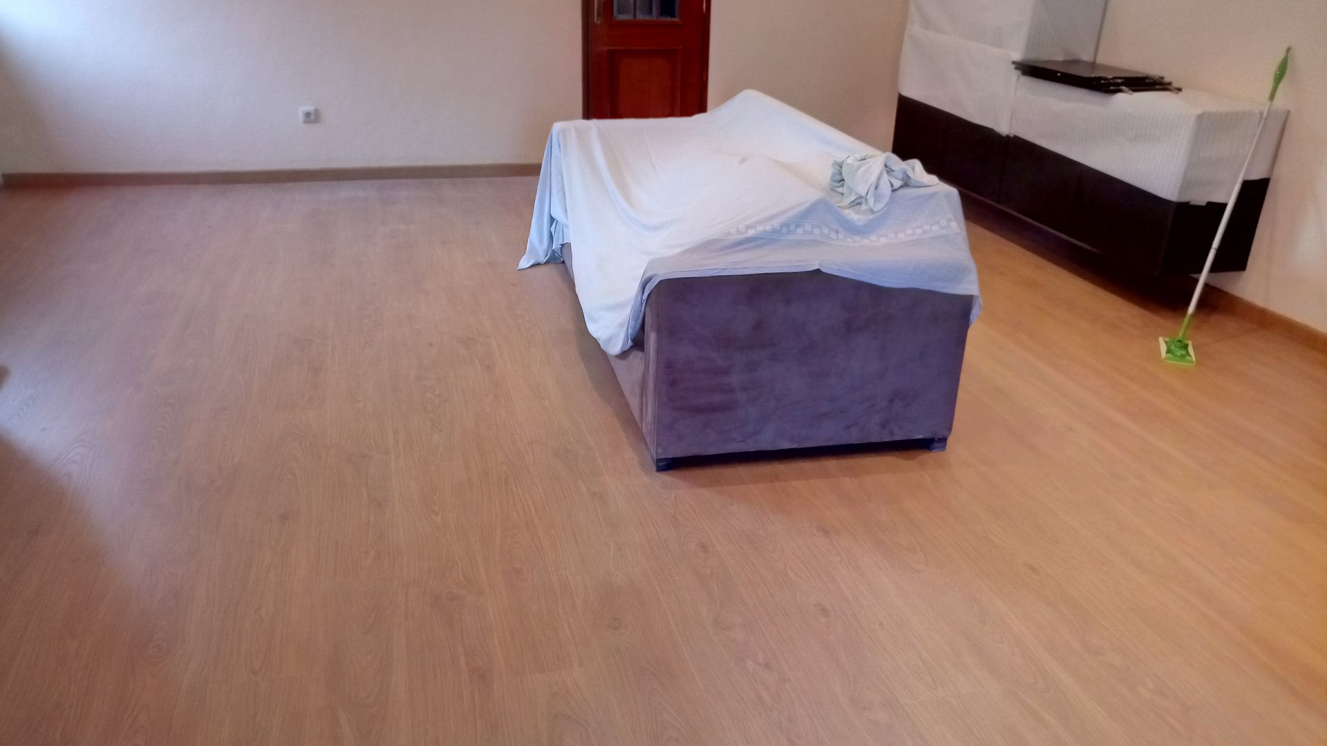 Quarto com piso de madeira, sofá coberto por um lençol branco, esfregão no canto e porta marrom.