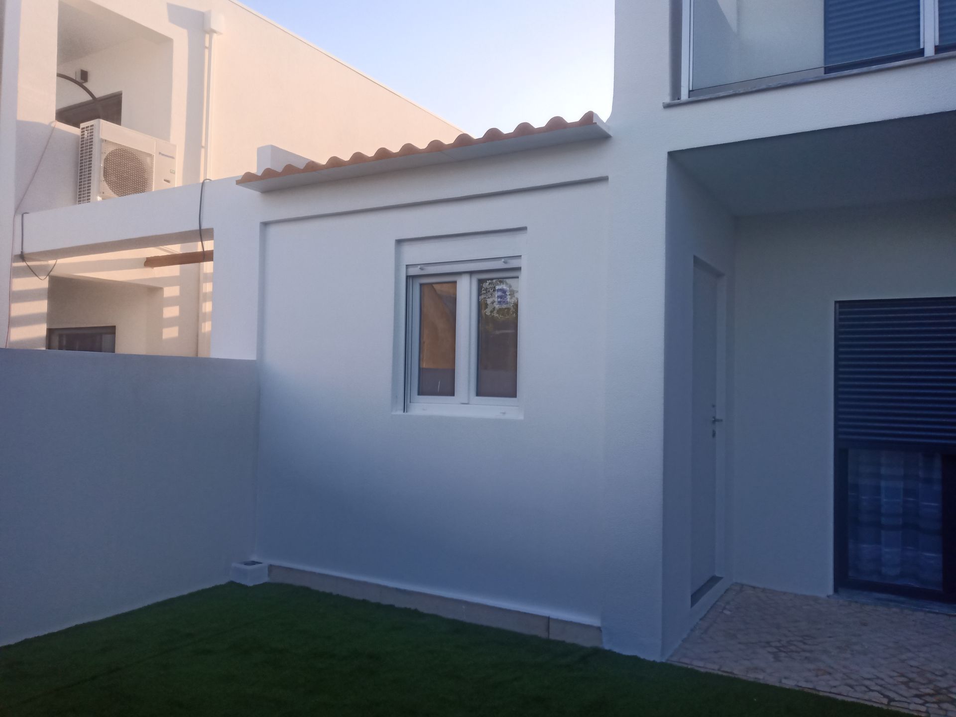 Exterior de um prédio branco com uma pequena janela, porta e telhado de telhas, ao lado de uma área gramada.