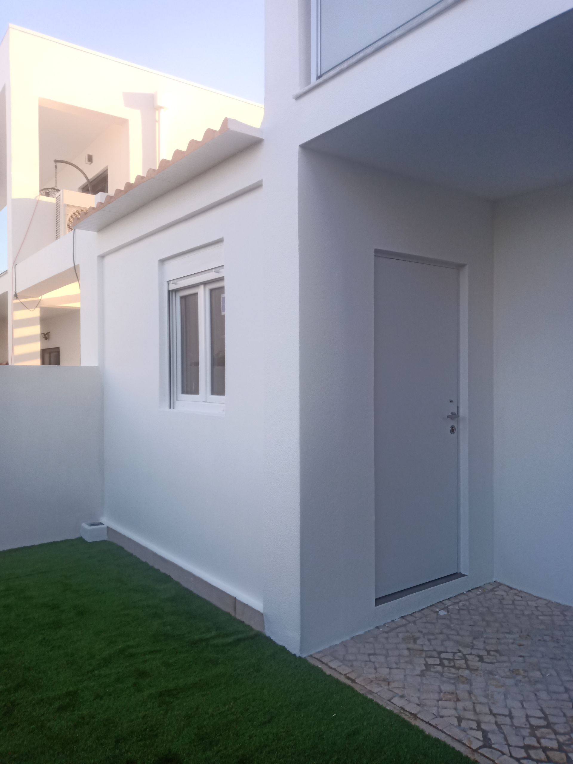 Exterior de prédio branco com porta, janela e uma pequena área gramada.