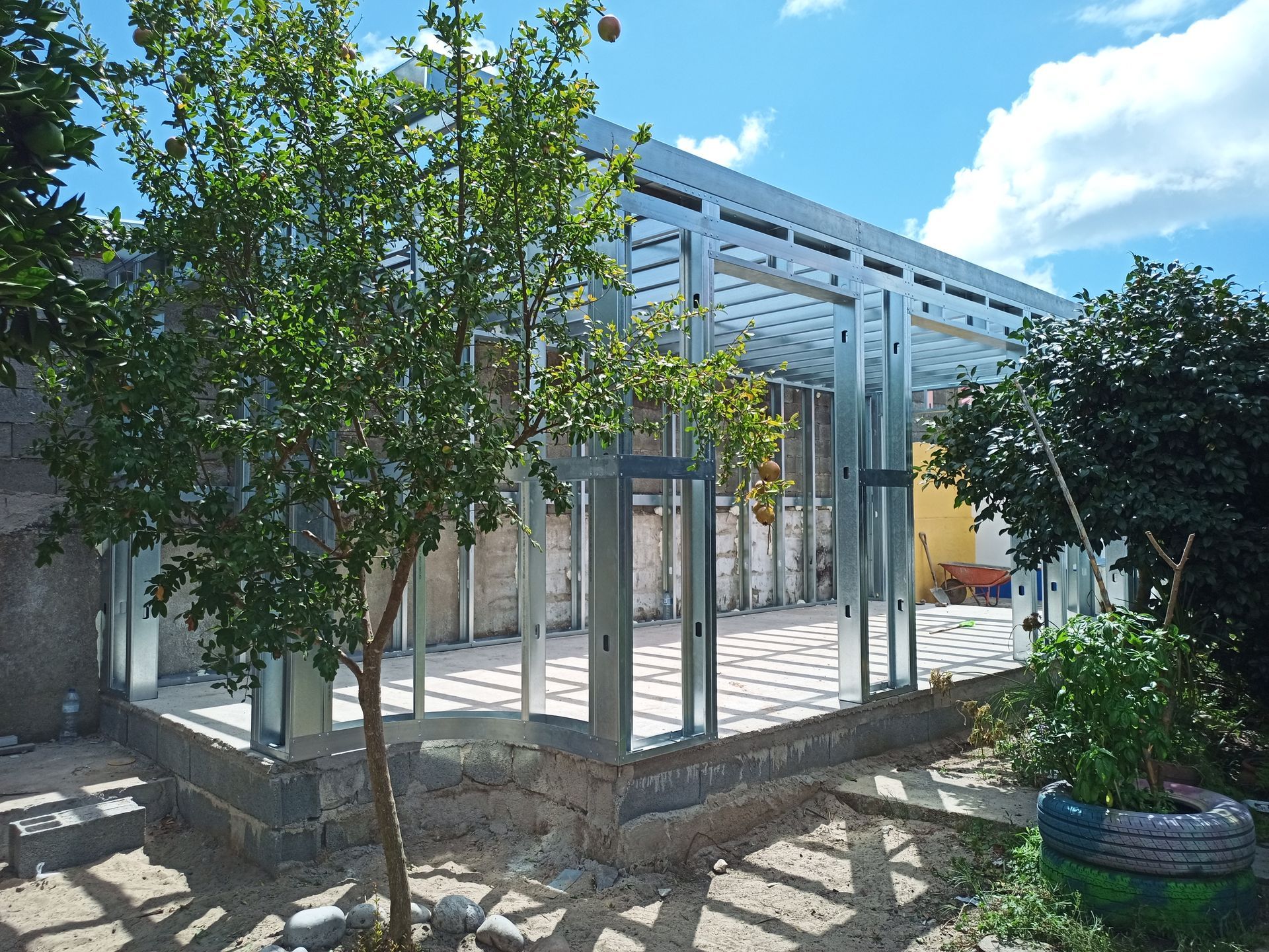 Estrutura metálica de um edifício em construção, assentada sobre uma fundação de blocos, com árvores e céu azul.