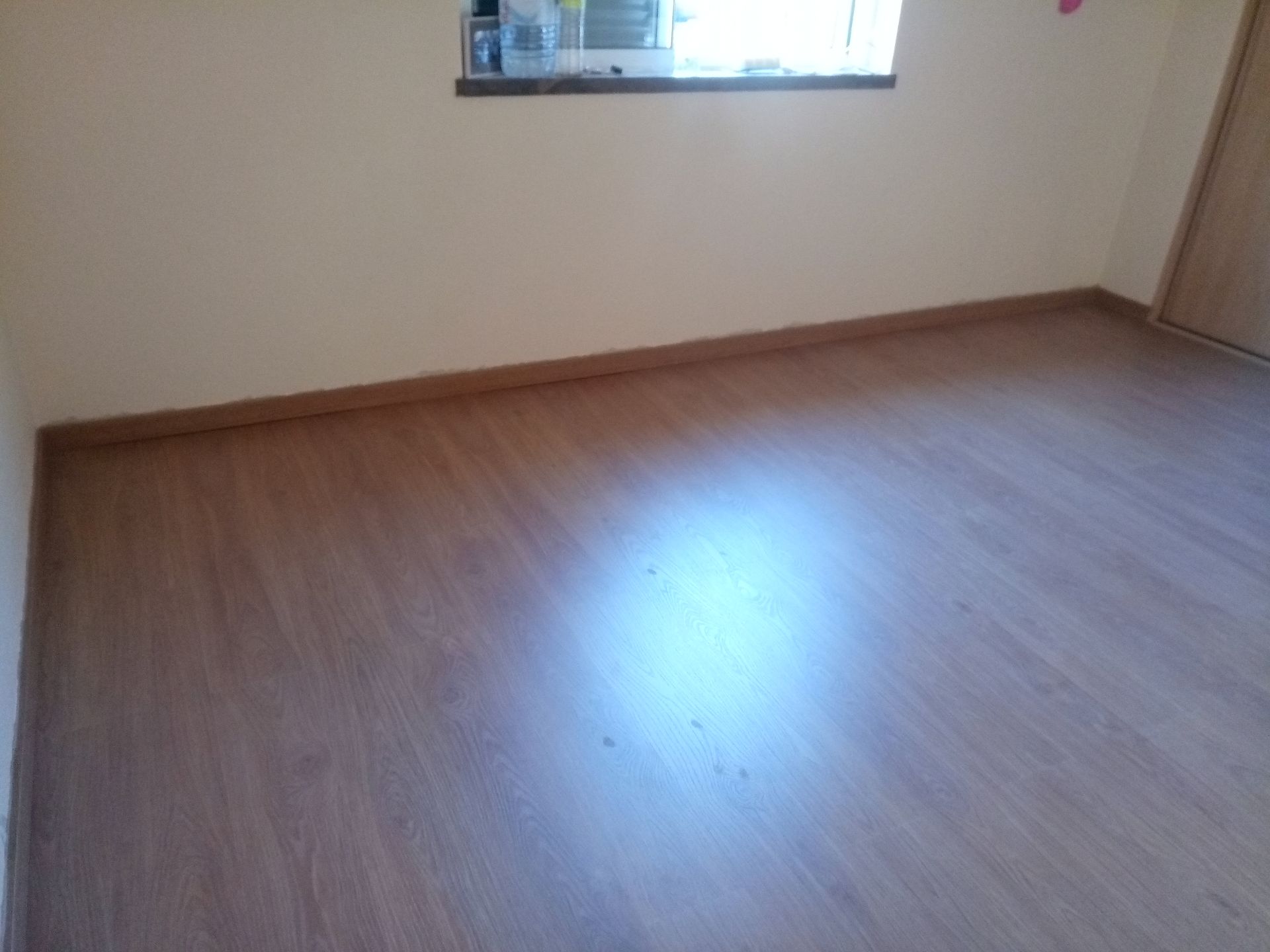 Quarto vazio com piso e rodapé imitando madeira. Janela na parede, porta à direita.