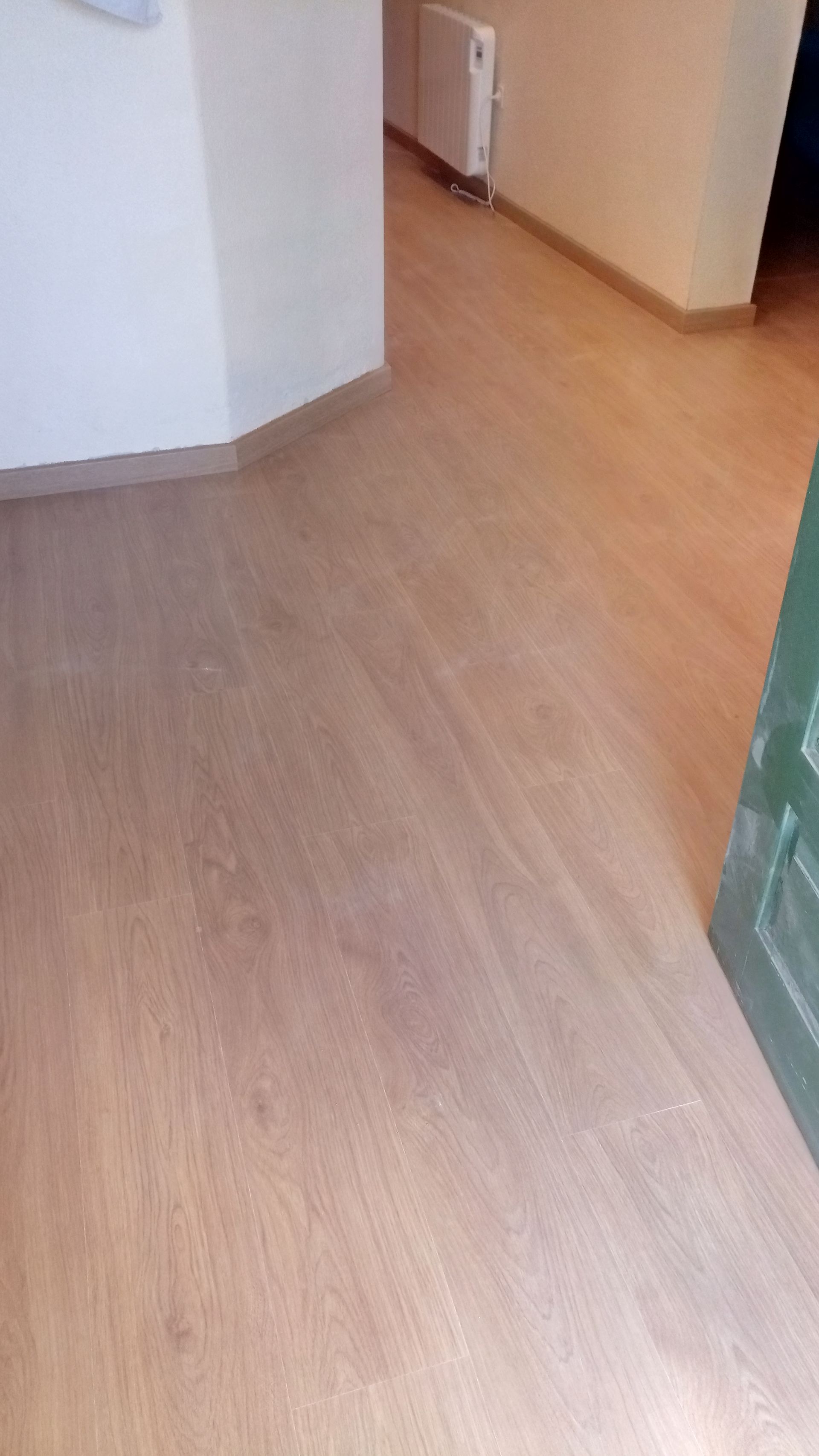 O piso que imita madeira em tom castanho claro se estende até o corredor. Uma parede branca e uma porta verde são visíveis.