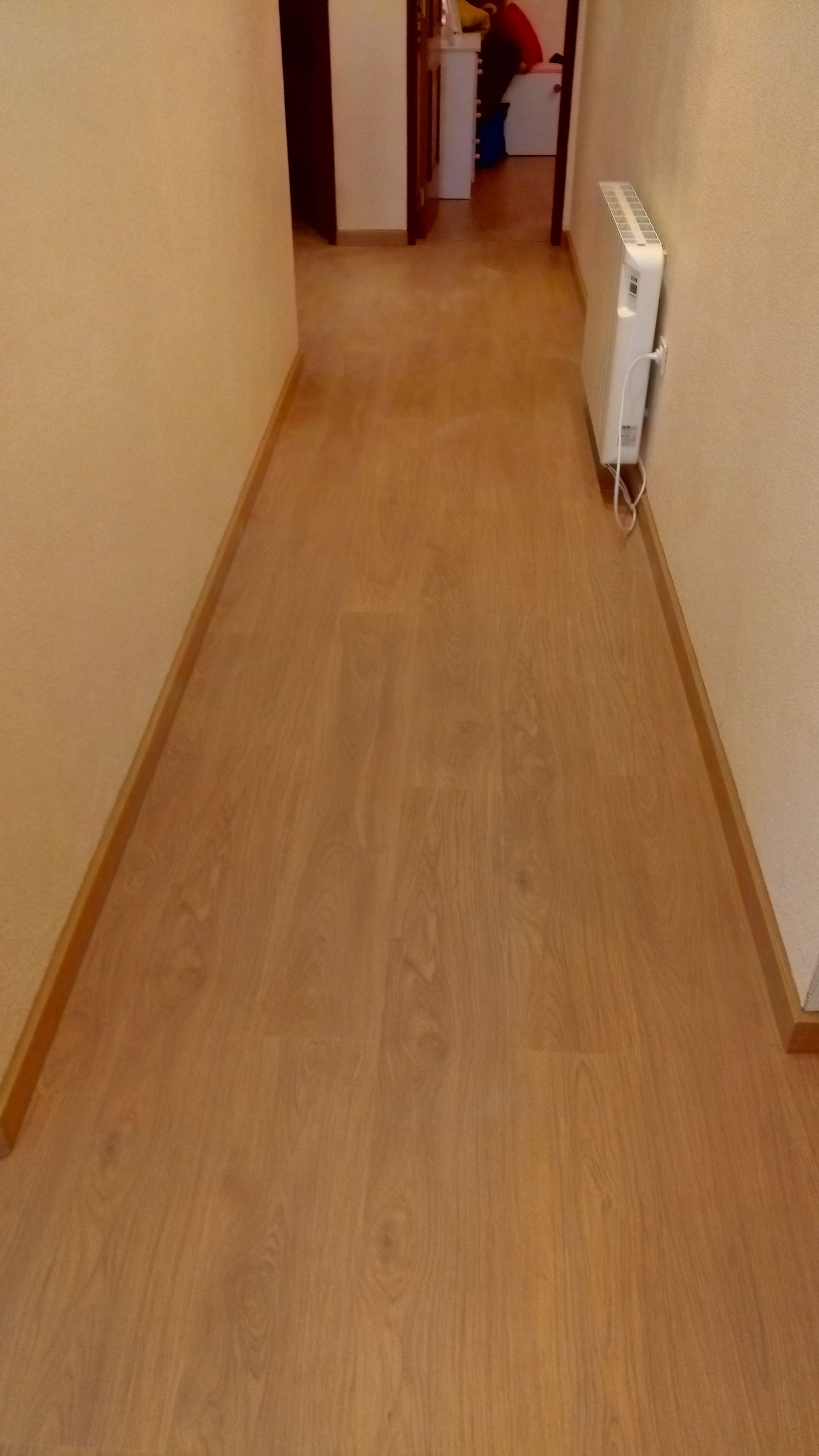 Corredor estreito com piso de madeira castanho-clara e paredes bege. Um radiador branco está instalado em uma das paredes.