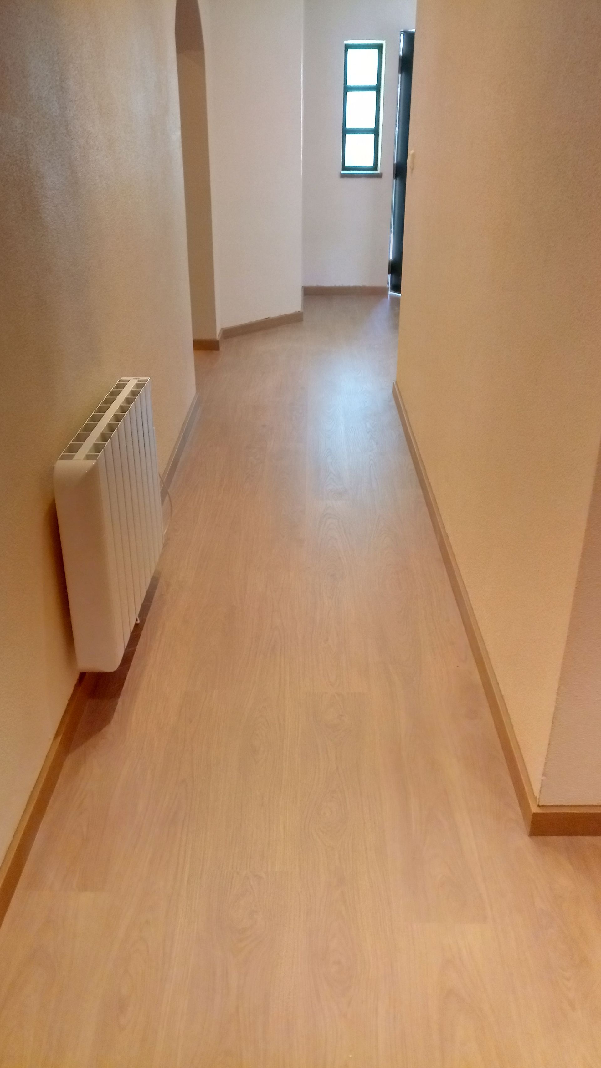 Corredor estreito com piso imitando madeira e paredes cor creme. Um radiador está instalado na parede esquerda.