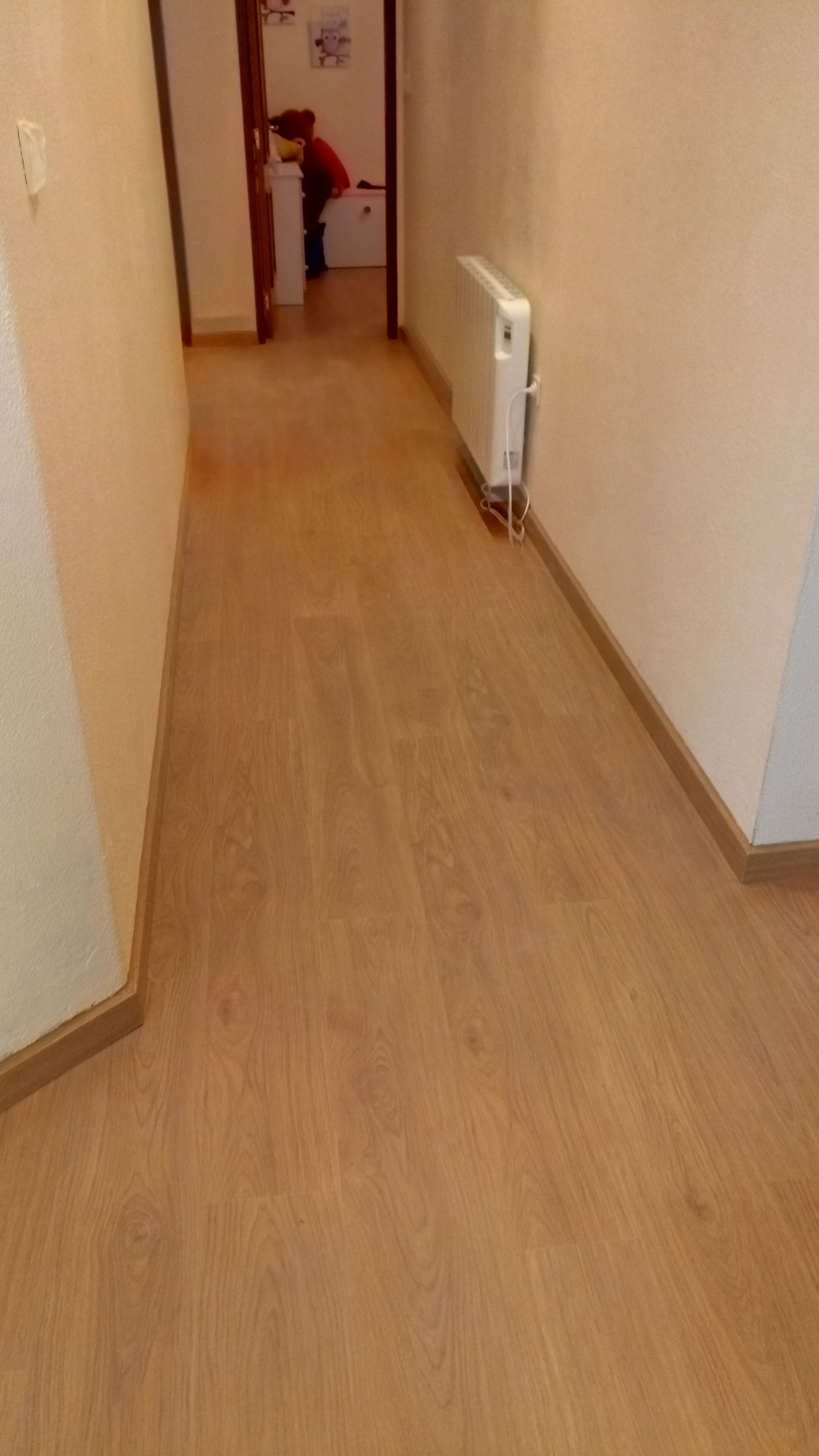 Corredor estreito com piso de madeira clara, paredes bege e um radiador. Uma pessoa é visível na porta.