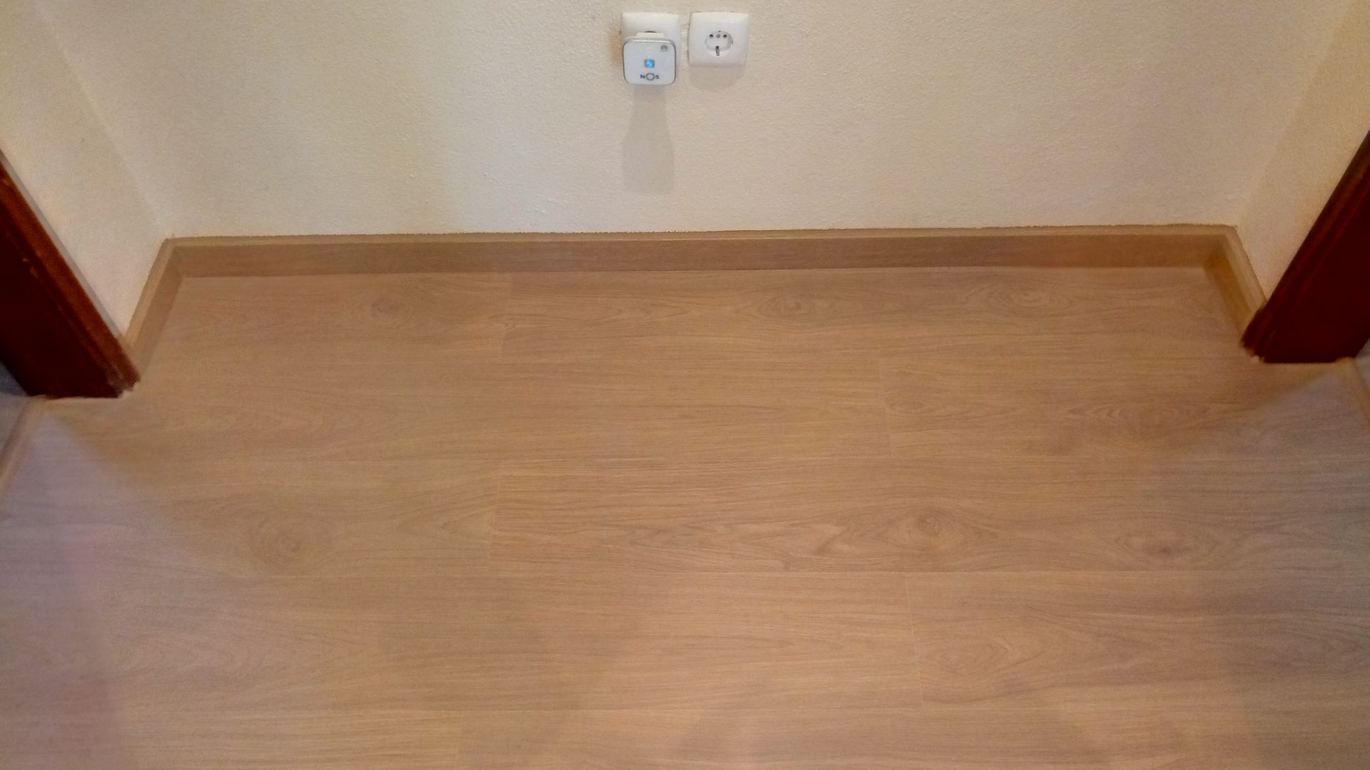 Piso e rodapé de madeira contra uma parede bege, com as molduras da porta visíveis, tomada e interruptor.