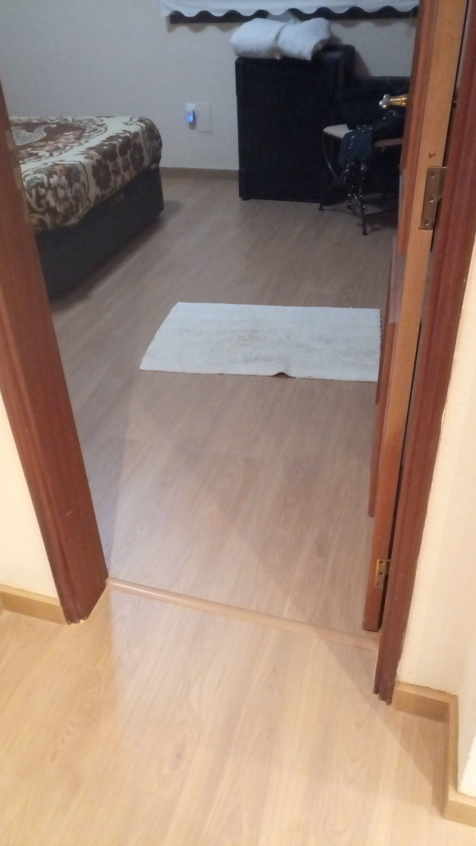 Interior de quarto com porta aberta, piso de madeira, cama, tapete e móveis escuros.