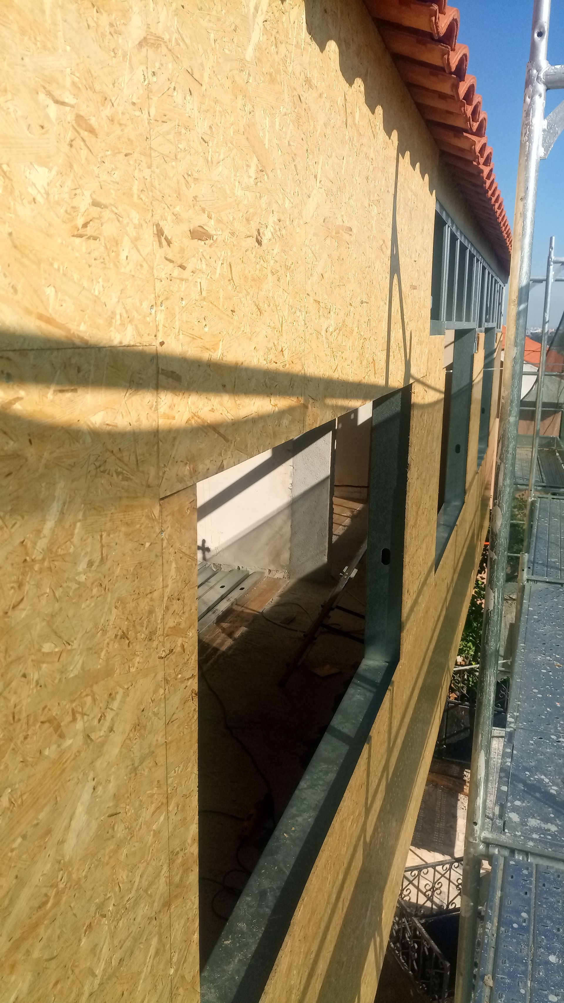 Canteiro de obras: Revestimento de OSB instalado ao redor da abertura de uma janela inacabada, com o telhado de telhas visível acima.