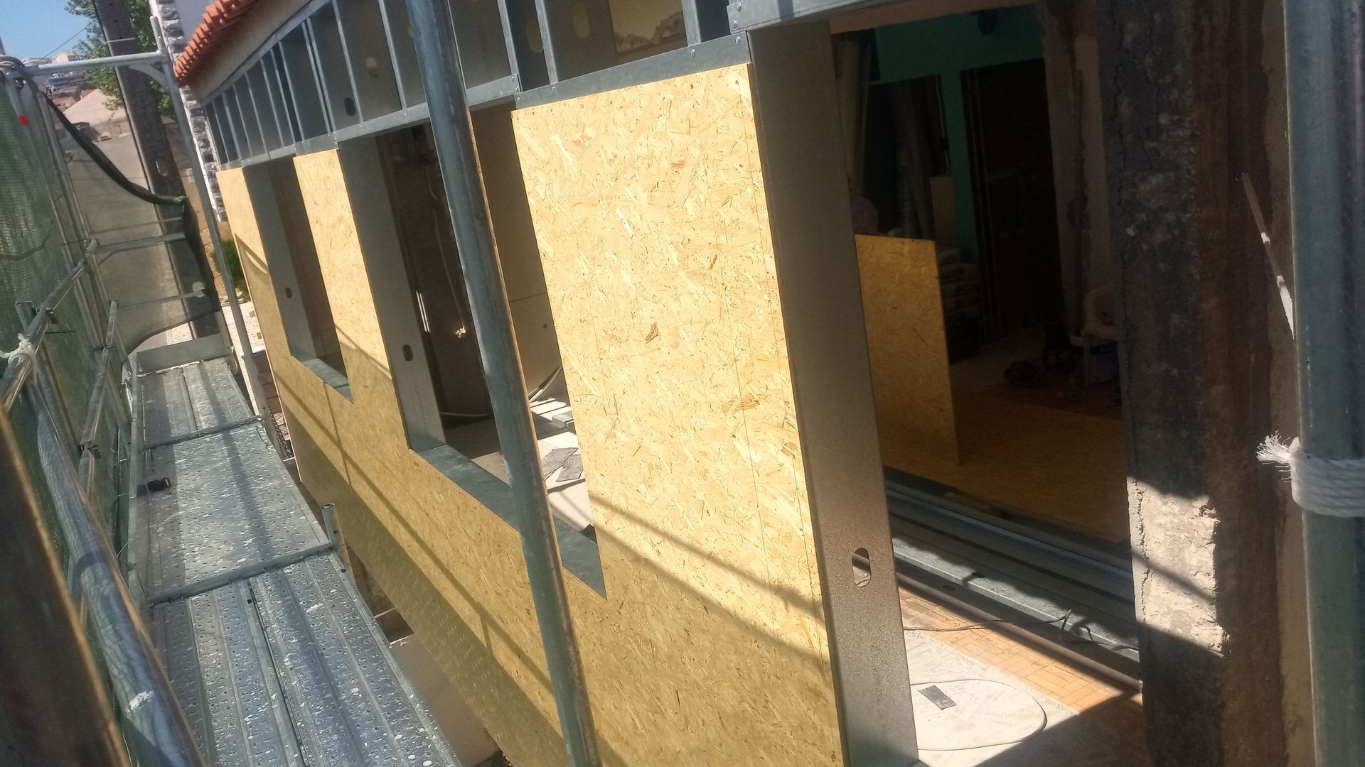 Vista externa do edifício em construção, mostrando painéis OSB, caixilhos de janelas e andaimes.