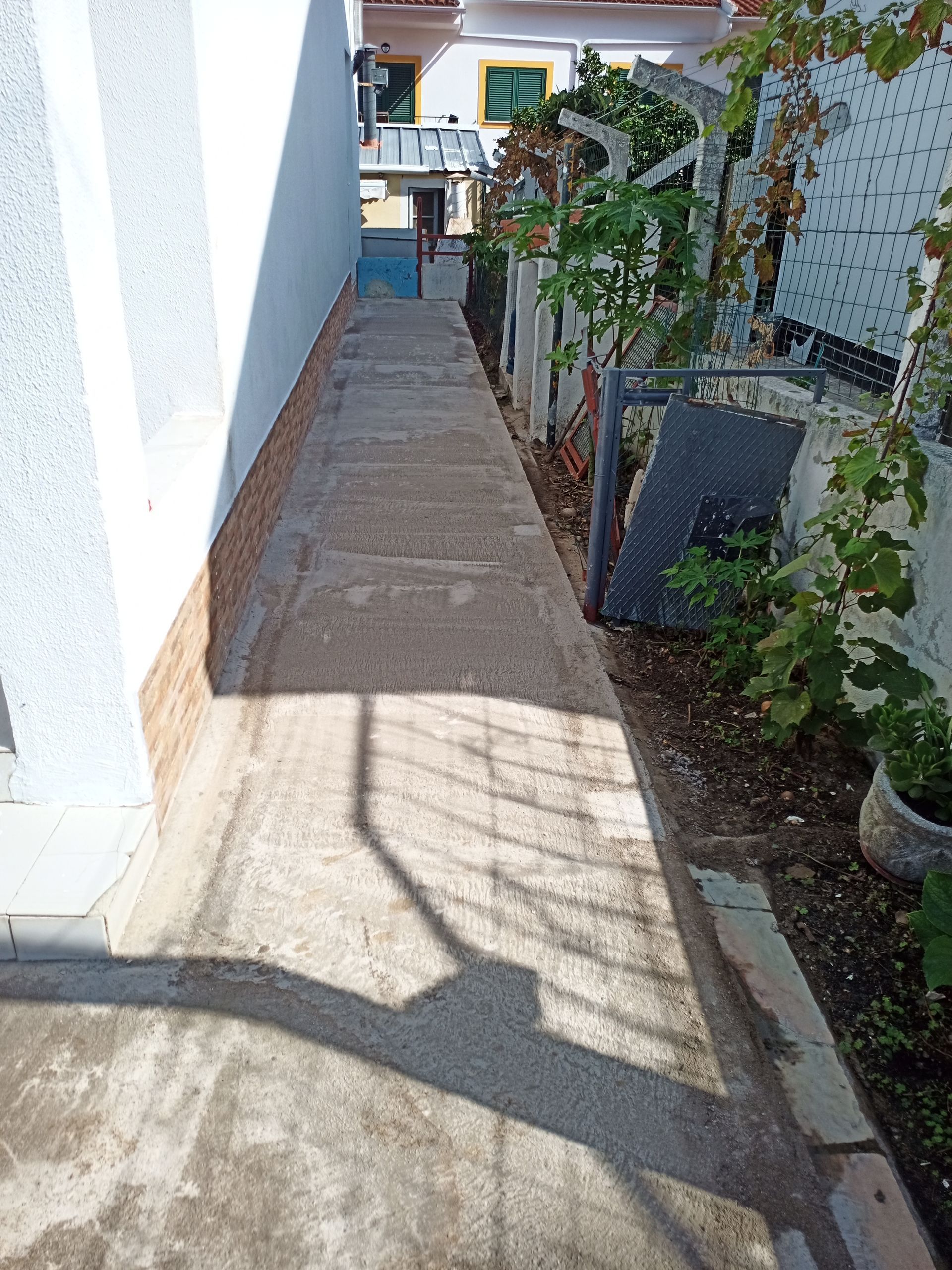 Caminho de concreto entre um prédio branco e uma cerca com plantas. Sombra de uma pessoa visível.