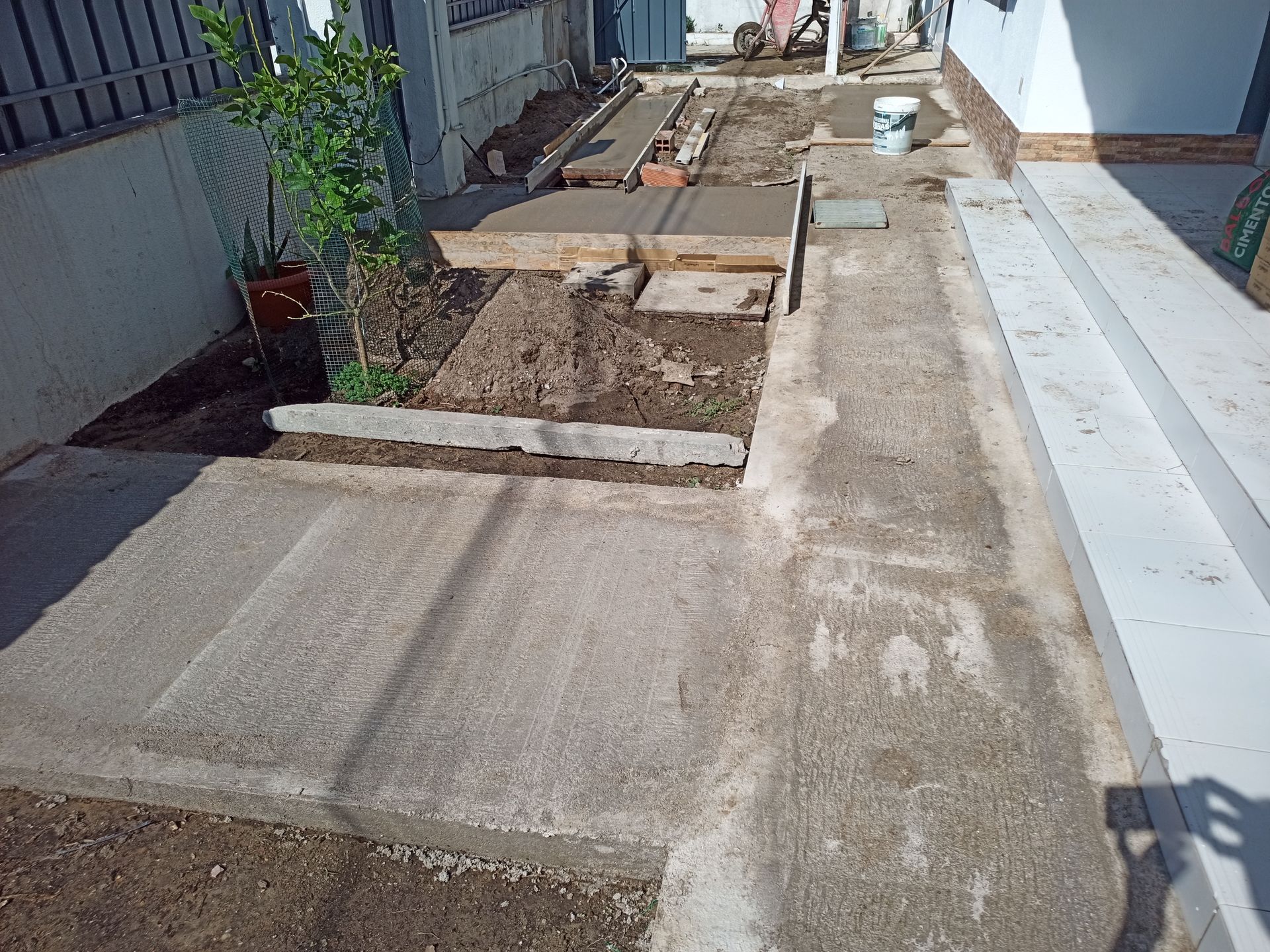 Obra de concreto em andamento, em ambiente externo. Calçada concluída e trechos inacabados, terra e plantas.