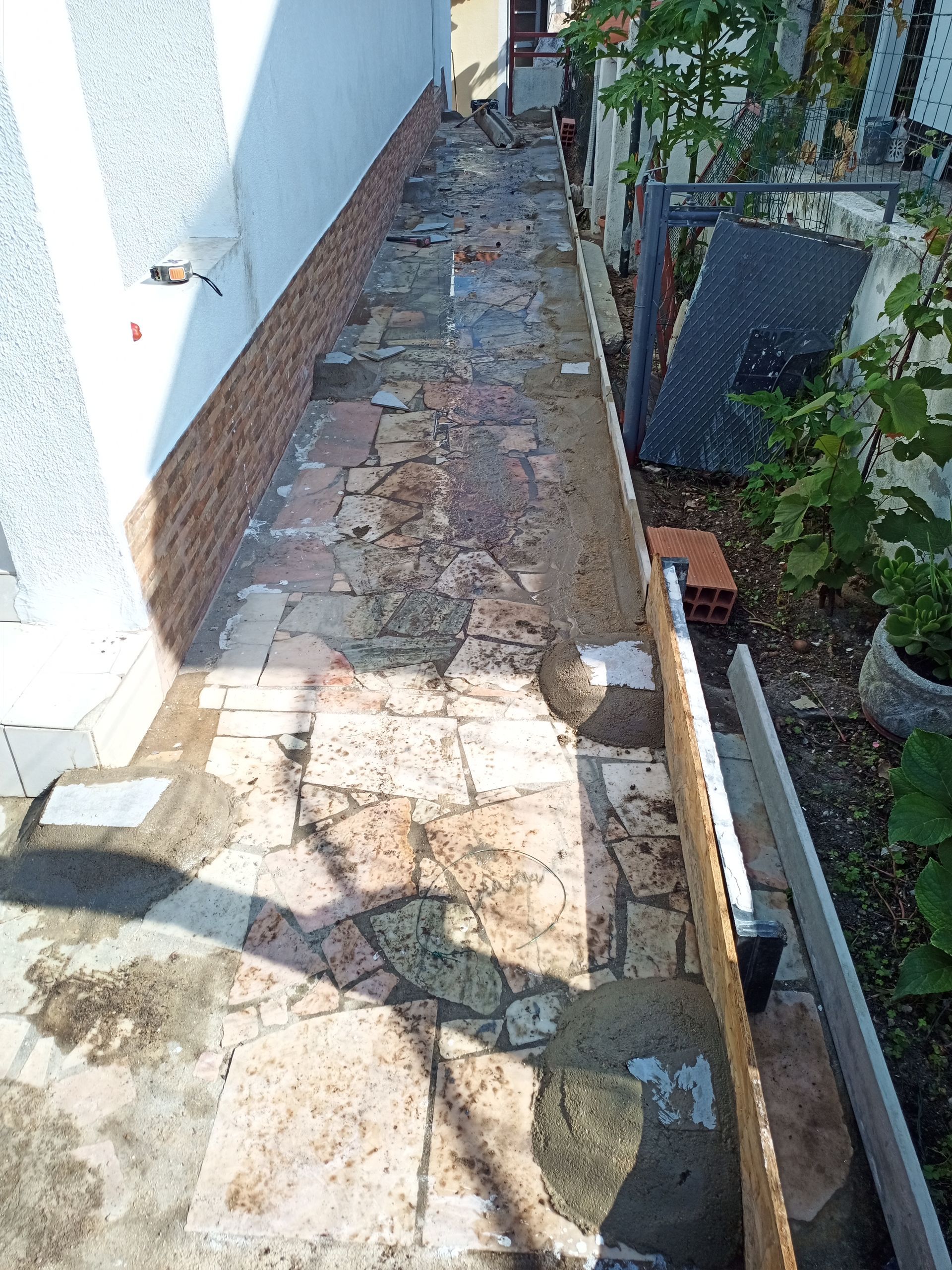 Caminho estreito pavimentado ao lado de um edifício. O pavimento de pedra está desgastado, com uma cerca de tijolo e metal à direita.