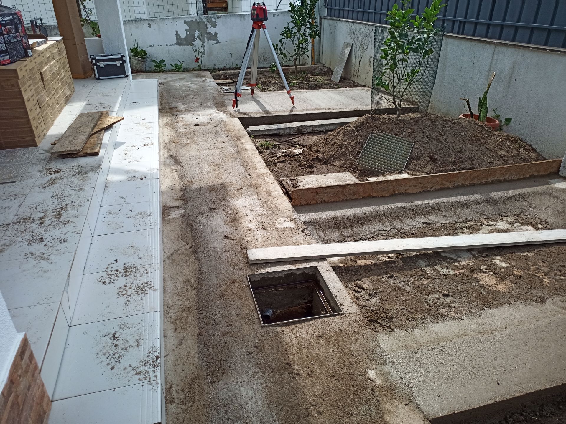 Canteiro de obras: Caminho de concreto com canteiros de terra expostos, grelha de drenagem, equipamento de topografia.