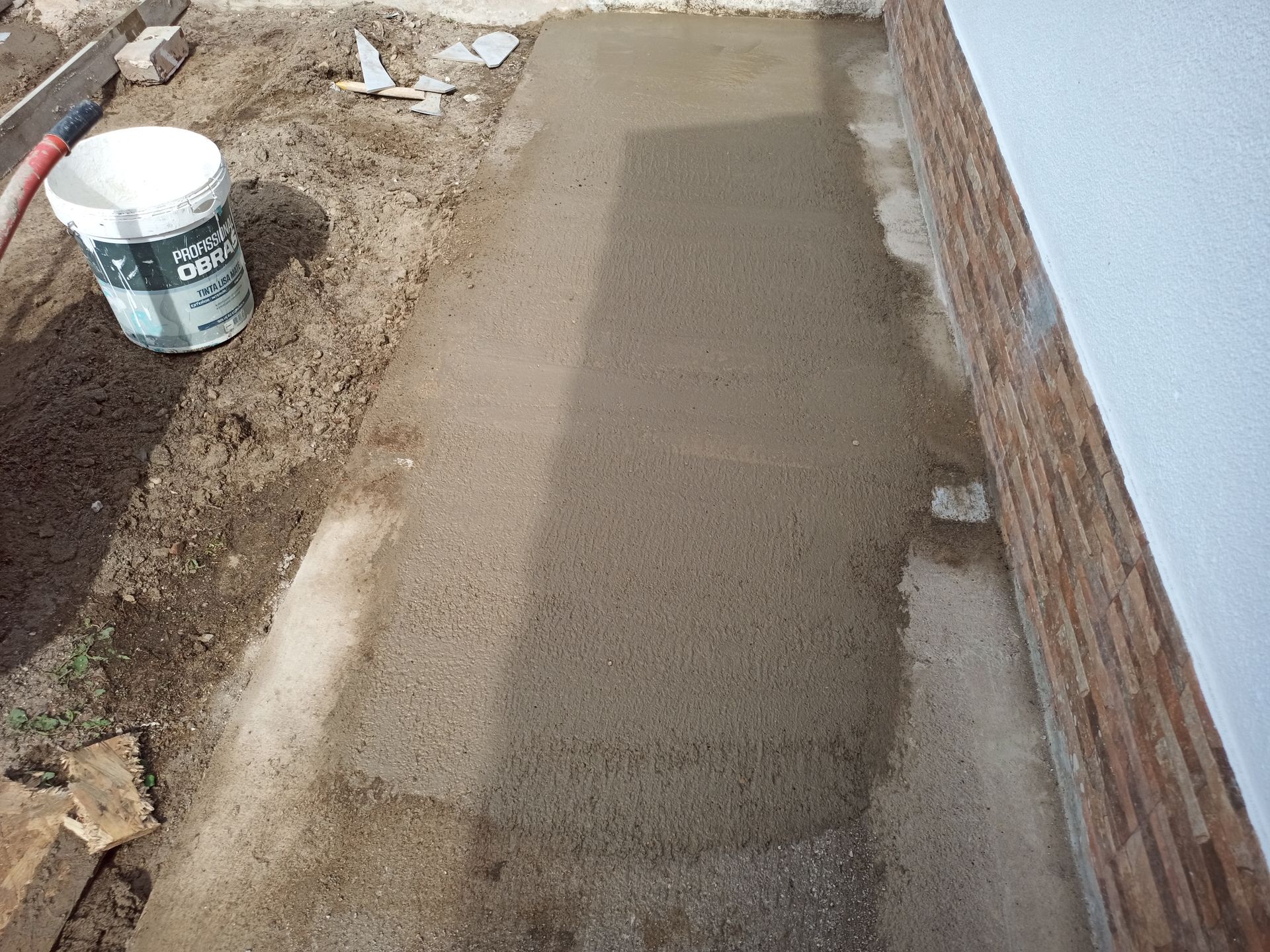 Caminho de concreto novo ao lado de uma parede de tijolos e uma parede branca, com um balde de tinta e sujeira visíveis.