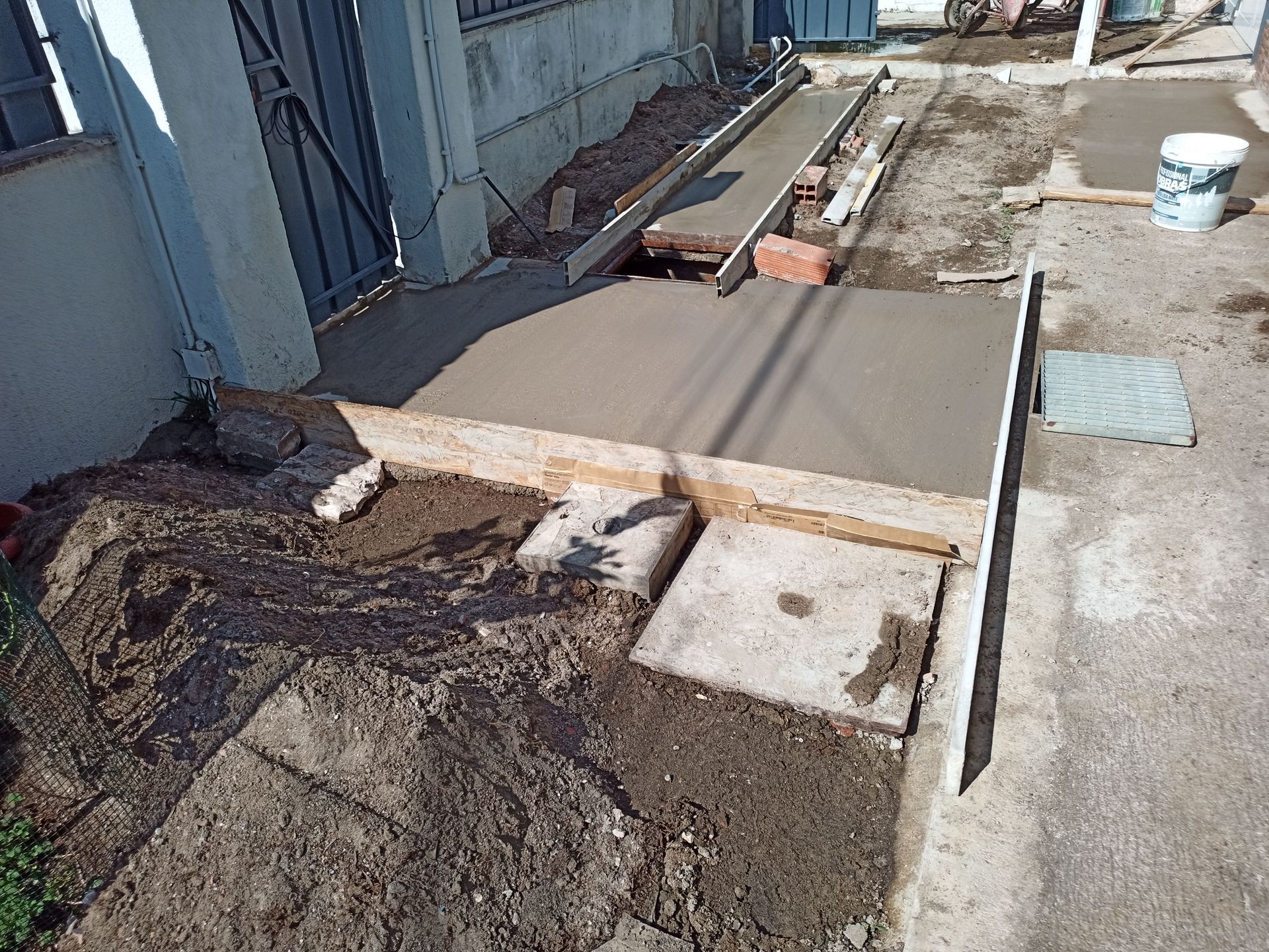 Construção de uma rampa de concreto na parte externa de um prédio; é possível ver o concreto fresco em fôrmas de madeira, ferramentas e um balde.
