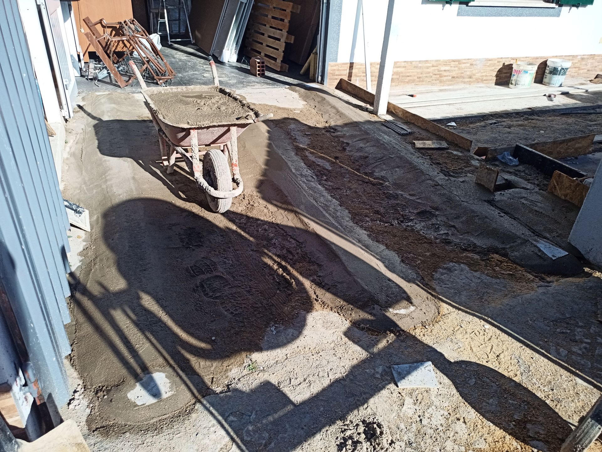 Carrinho de mão em uma entrada de concreto em construção, com sombras projetadas pelo sol.