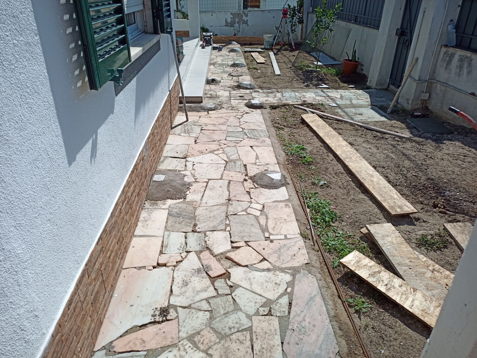 Caminho de pedra em construção ao lado do prédio, com paisagismo e tábuas de madeira.