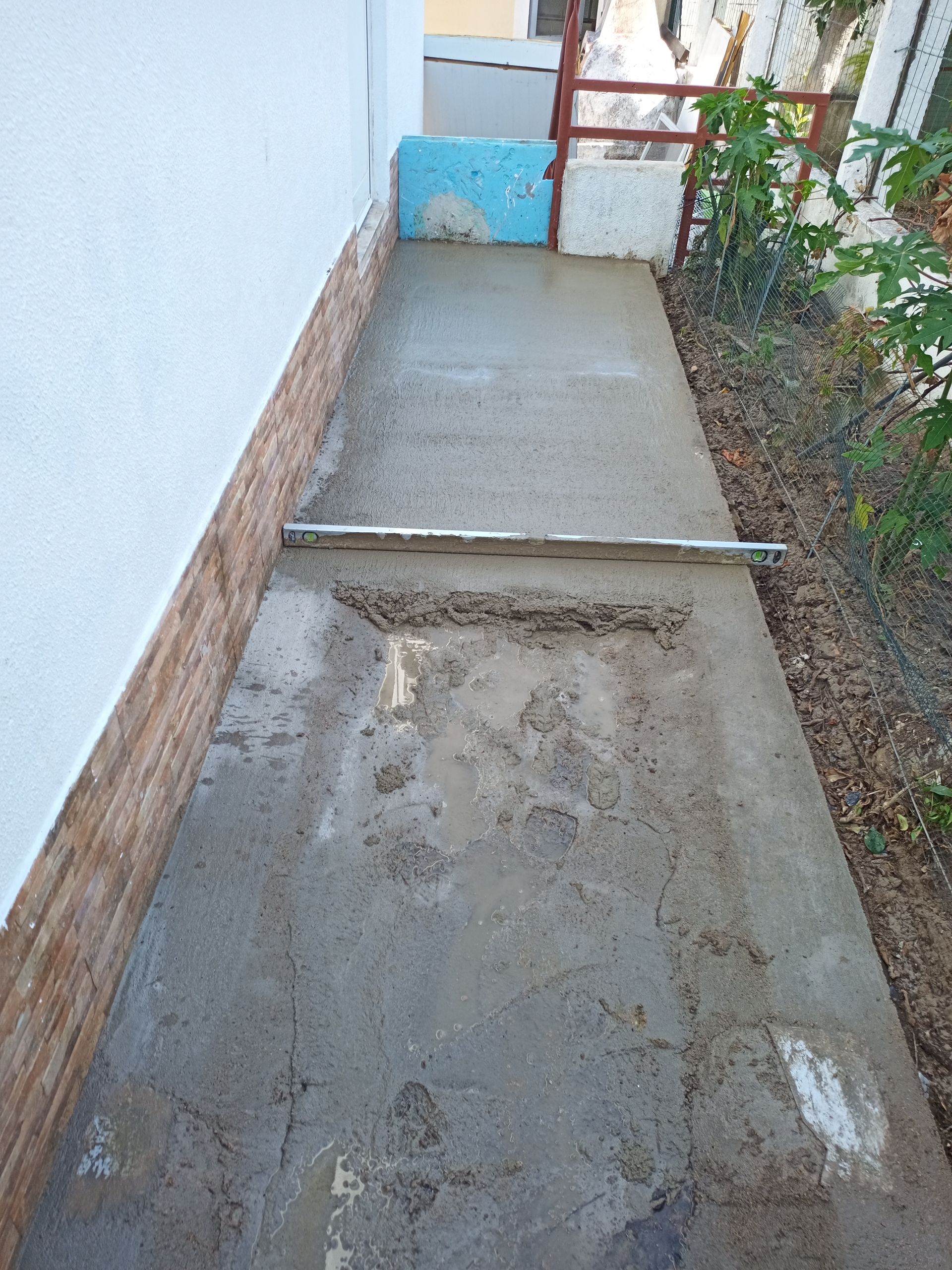Caminho de concreto ao lado de uma parede branca e um pedaço de terra com folhagem. Um nível está apoiado no concreto.