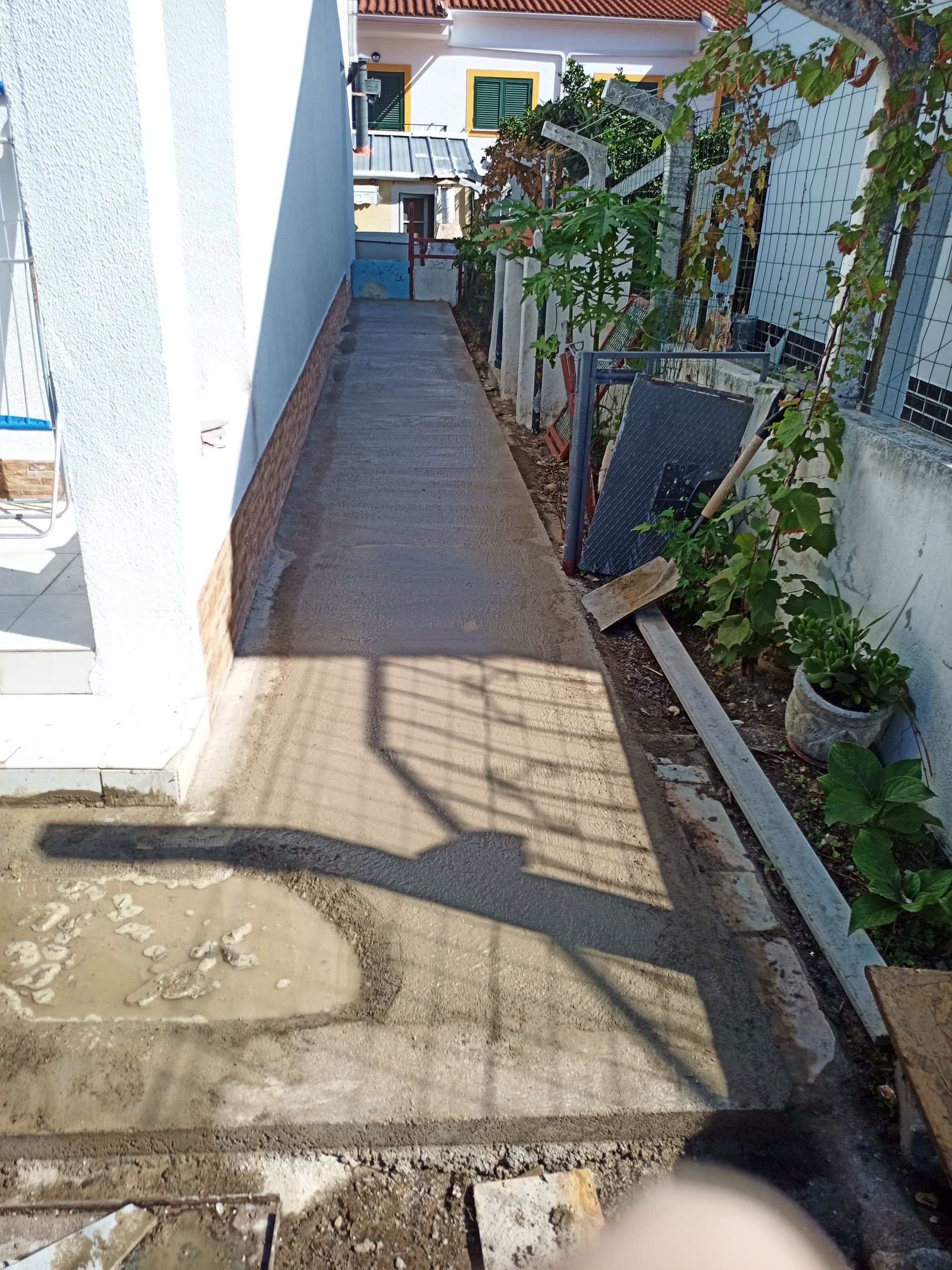 Passarela estreita de concreto entre um prédio branco e um muro com plantas, que leva a um portão.