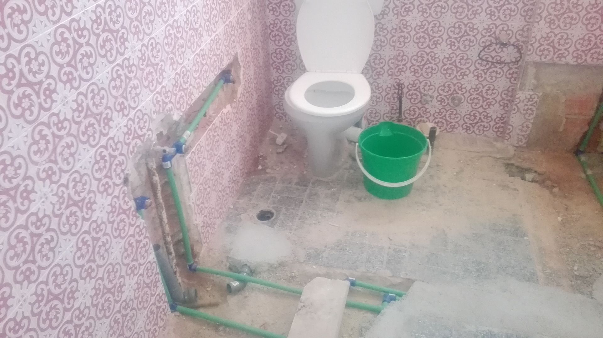 Banheiro com canos aparentes, vaso sanitário, balde verde e azulejos rosa estampados.
