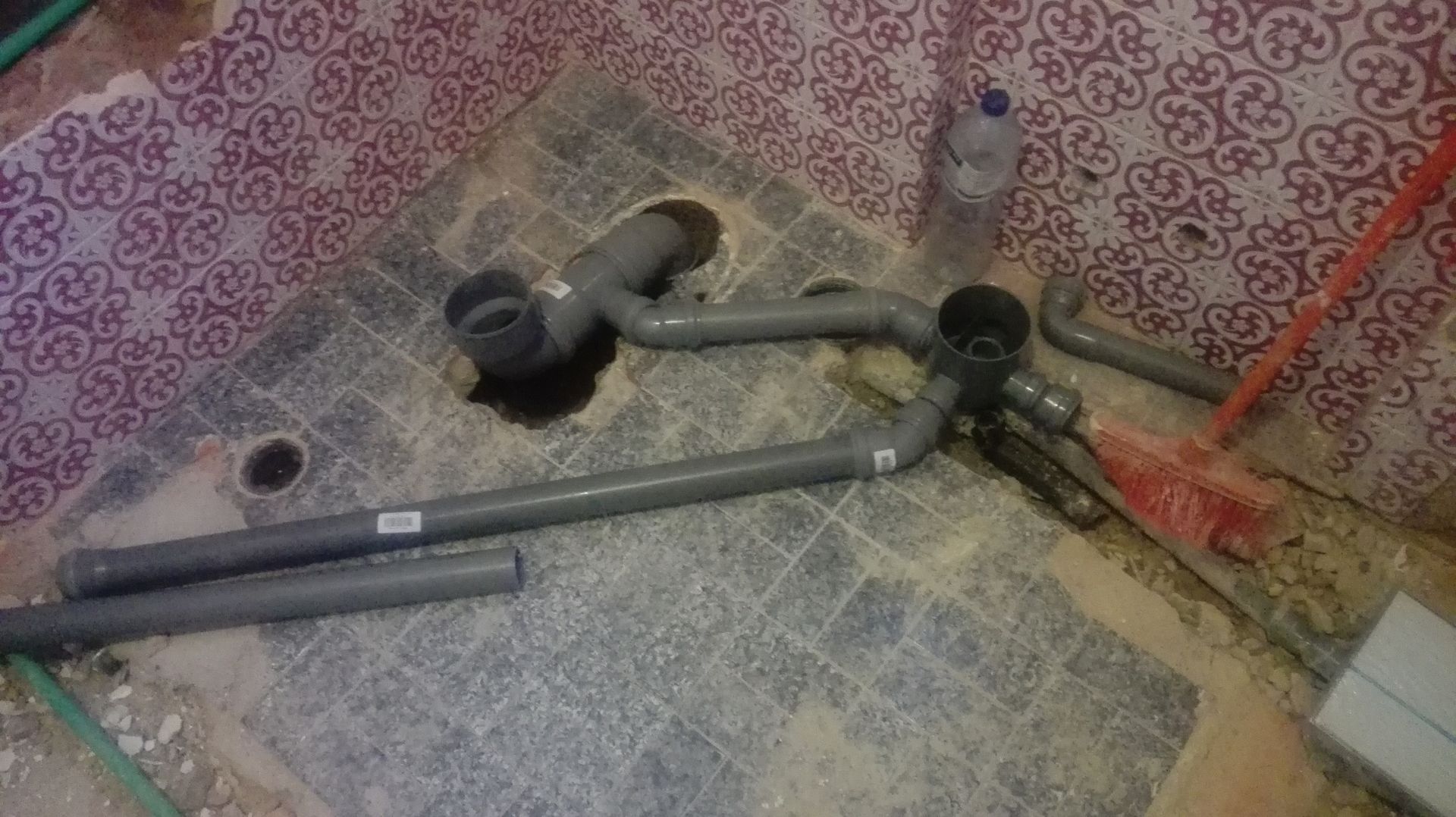 Tubos de PVC cinza e um ralo instalados sobre um piso de azulejos com paredes estampadas em rosa.