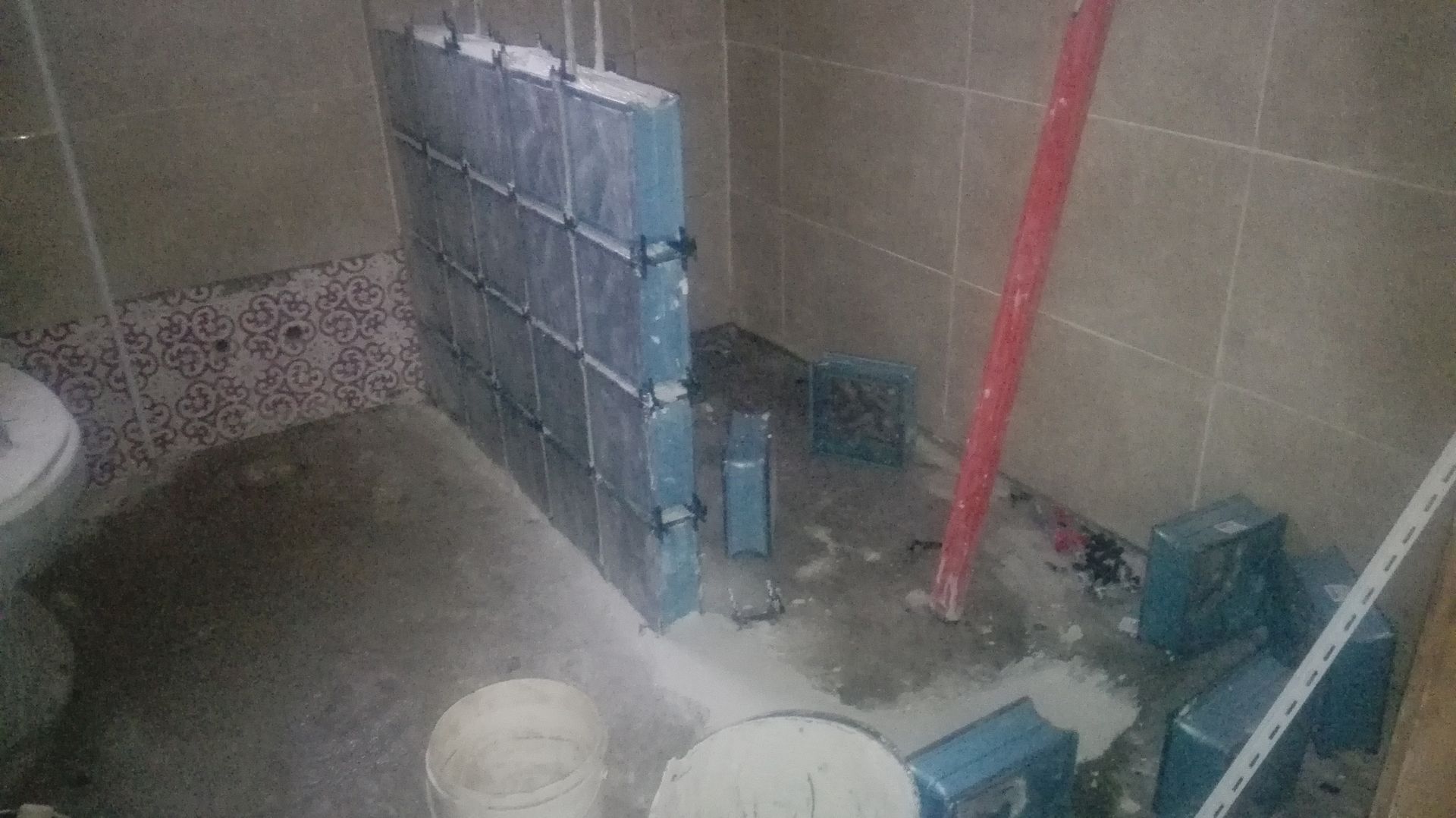 Banheiro em construção; azulejos azuis parcialmente instalados em uma parede. Piso de concreto e entulho.
