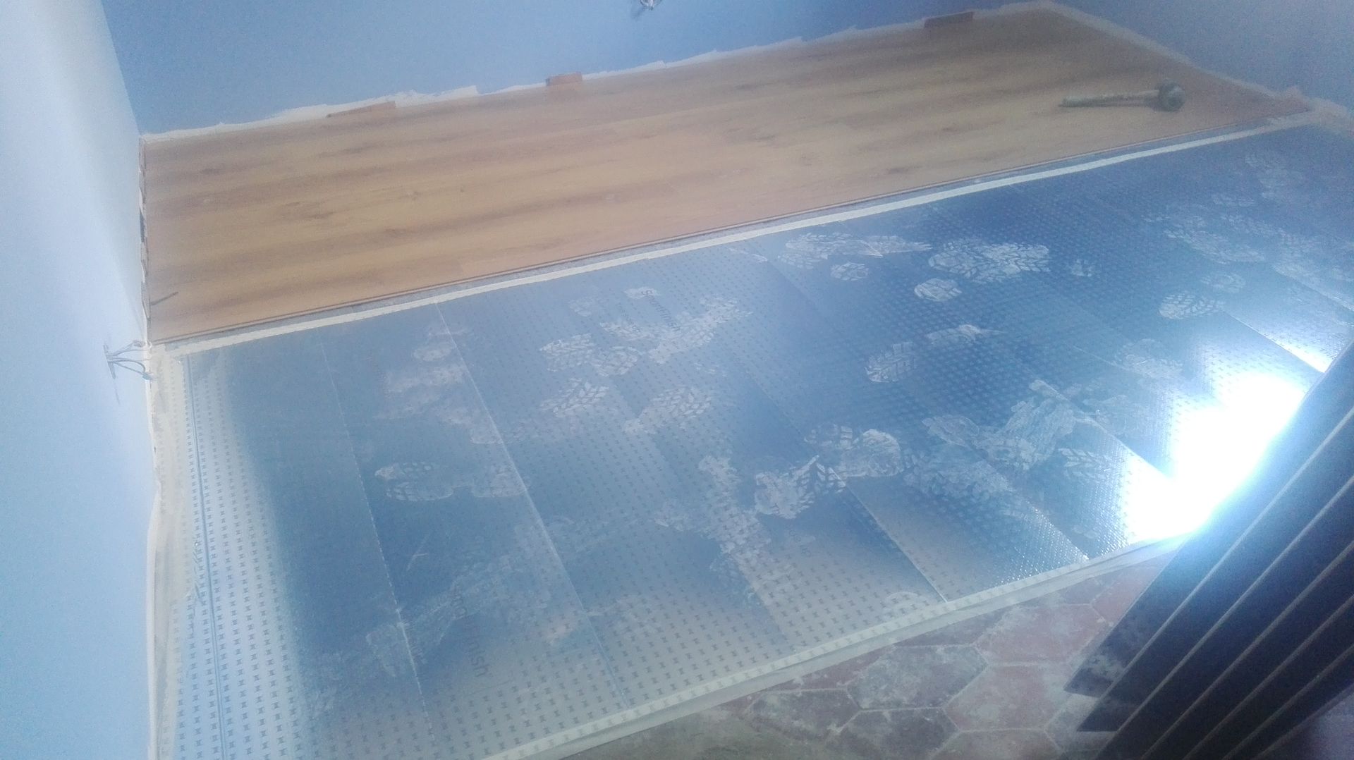 Piso com azulejos novos instalados ao lado de tábuas de madeira, coberto com plástico protetor.